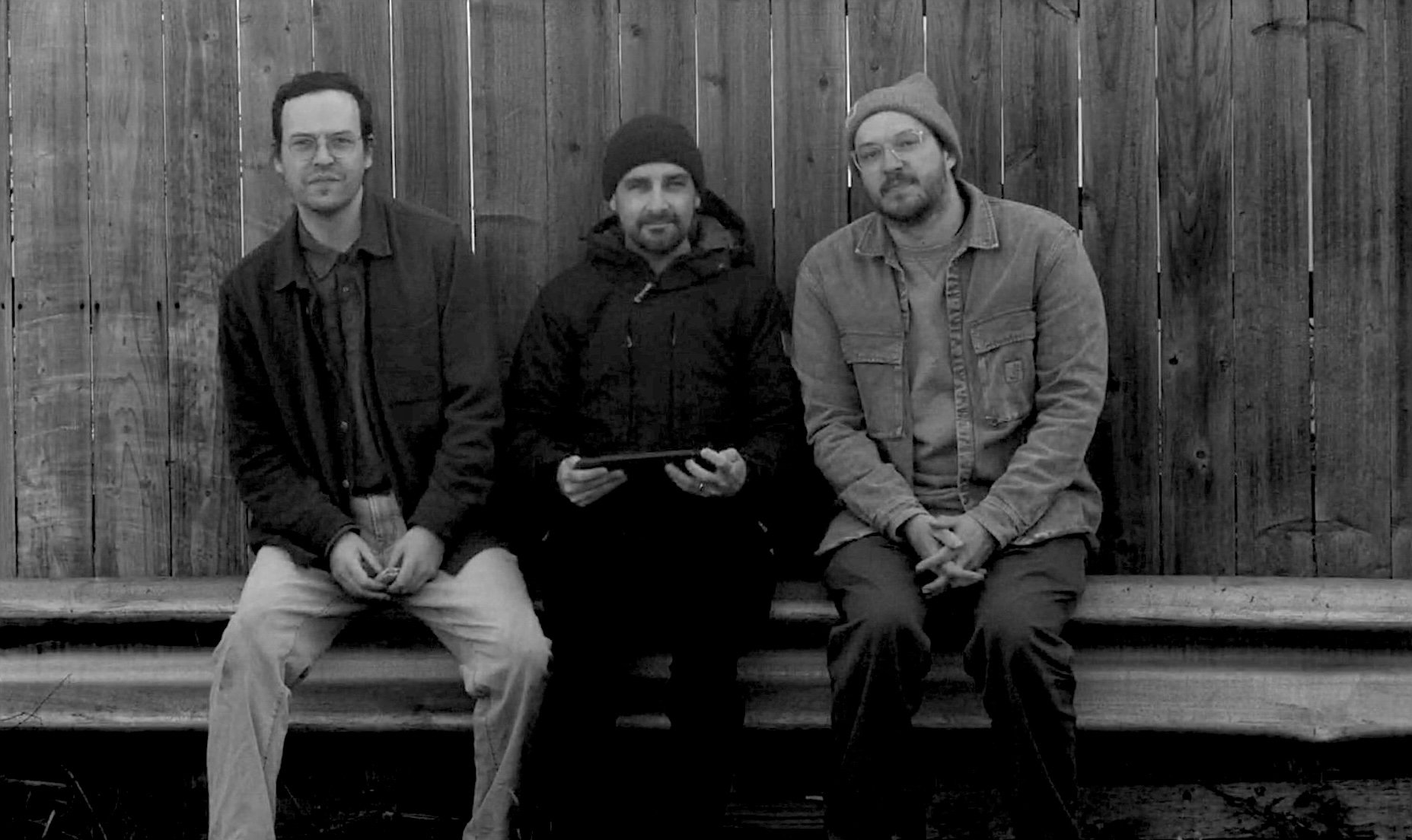 Three men sitting on a wooden bench outside in front of a wooden fence, dressed casually, with the middle man holding a tablet. John Anderson Beavers, cinematographer, director of photography
