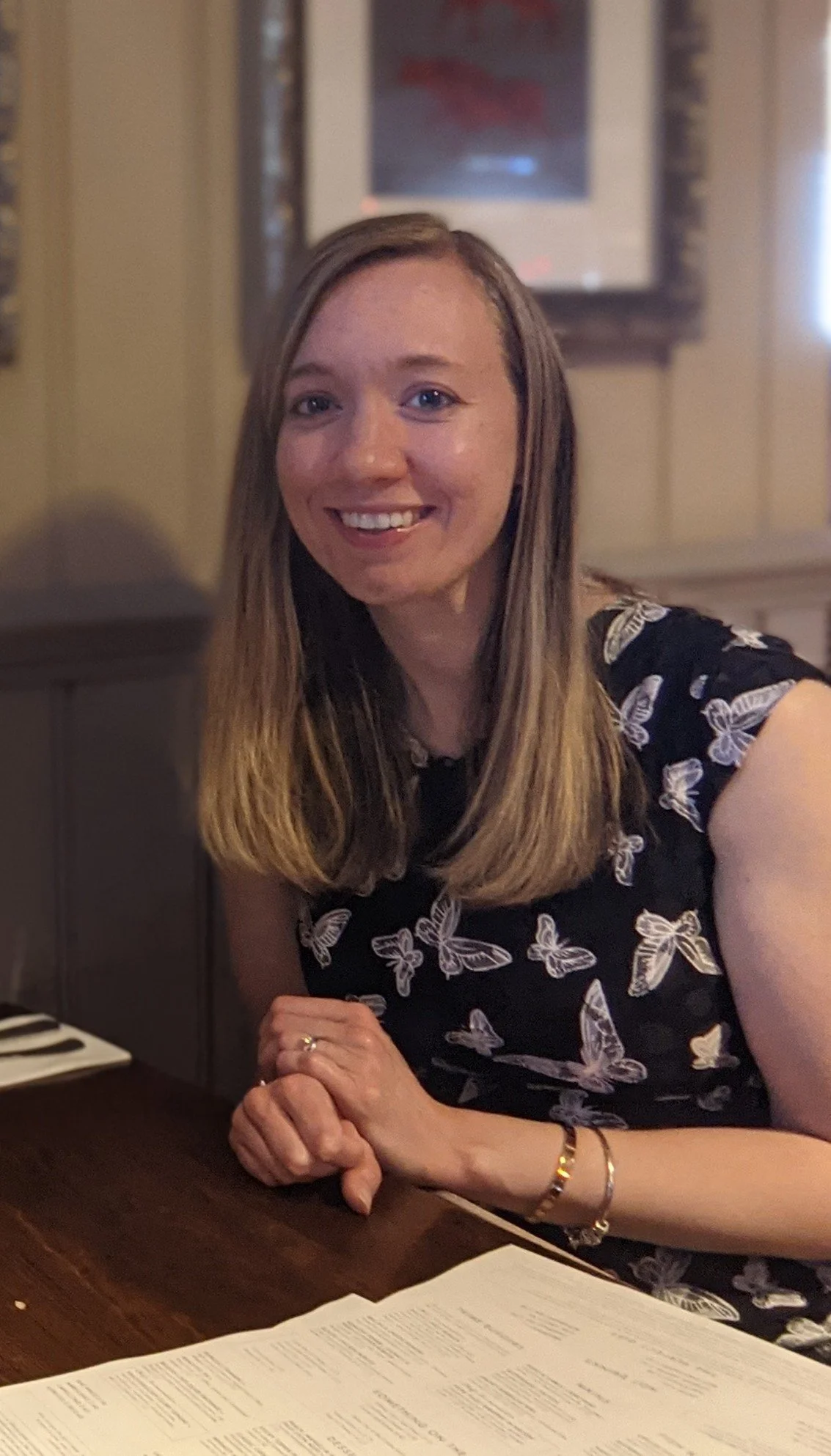 A woman in her 30s sitting at a table smiling at the camera. She has long blonde hair, blue eyes and is wearing a black dress detailed with white butterflies.