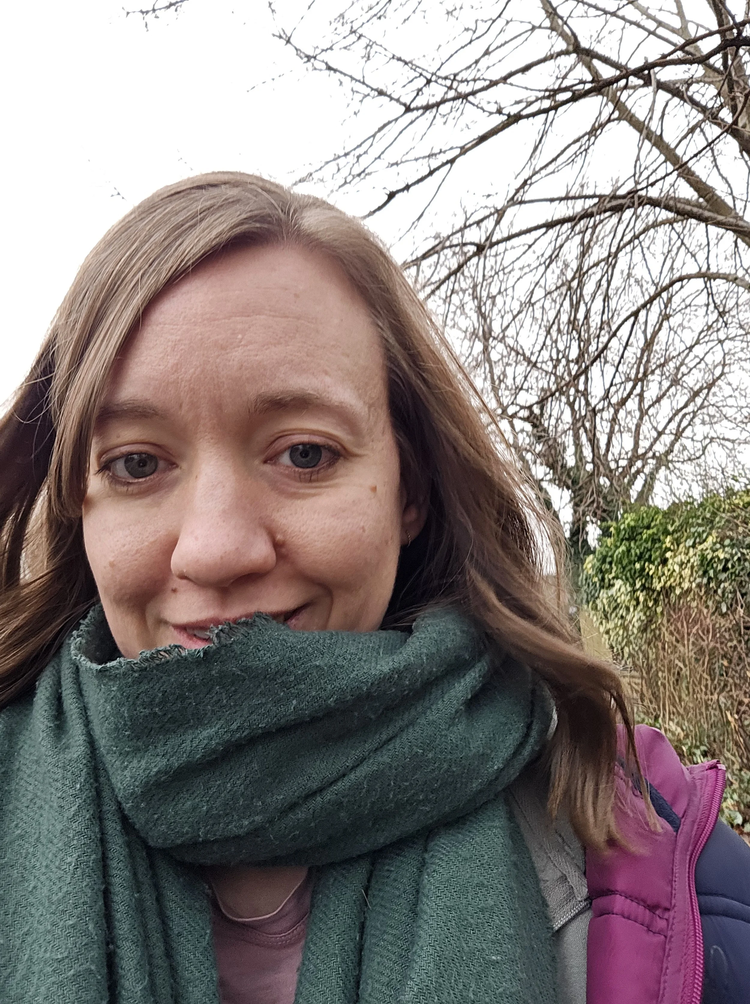 A woman with light brown hair, wearing a green scarf, pink jacket, and purple backpack, standing outdoors with leafless trees and some green foliage in the background.