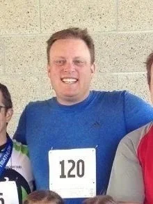A man wearing a blue shirt with a bib number 120 at an outdoor event, smiling and standing with other people.