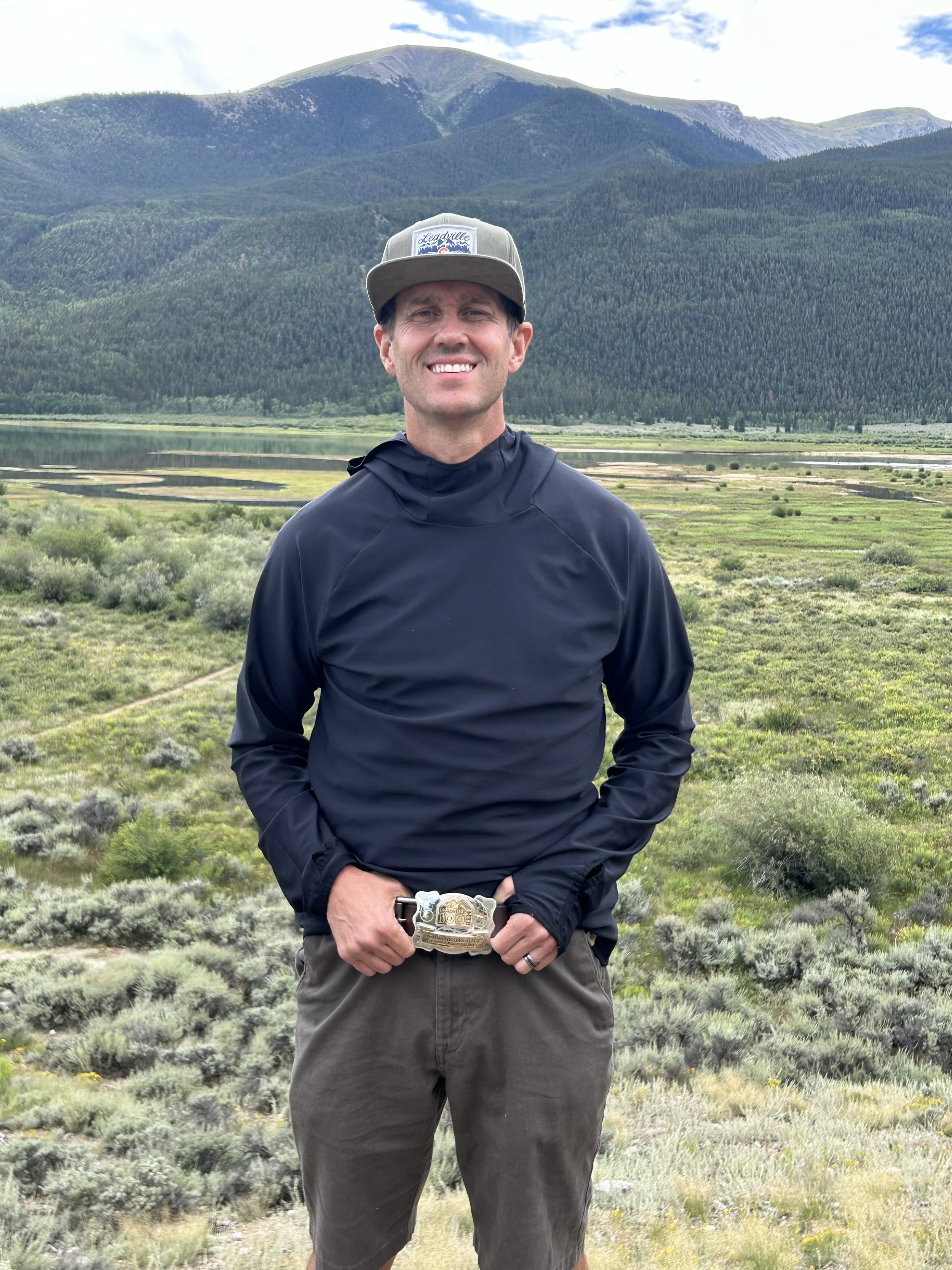 A man standing outdoors in front of a mountainous landscape with green terrain, wearing a black jacket, brown shorts, a gray hat, and a belt with a decorative buckle, smiling at the camera.