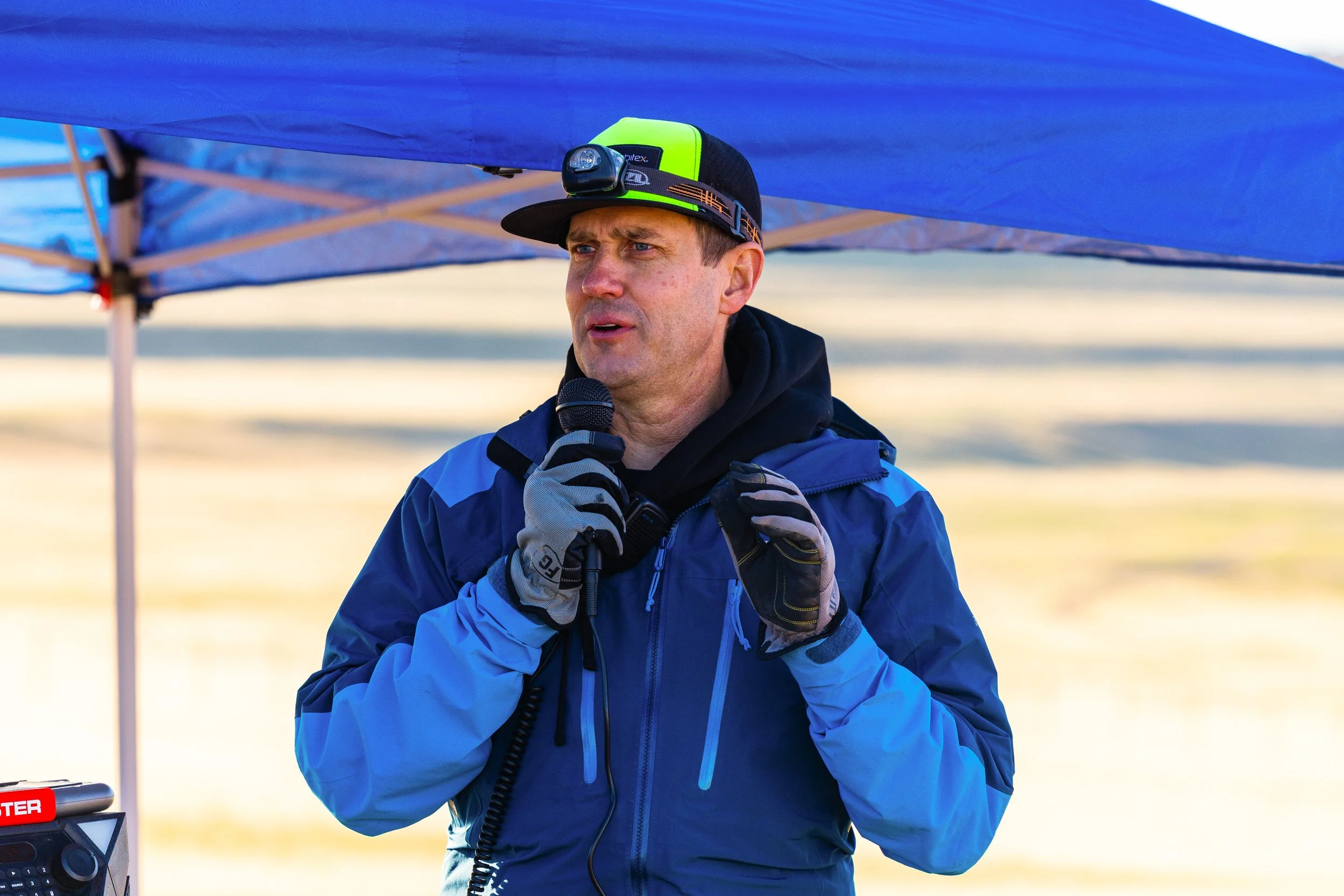 A man speaking into a microphone under a blue canopy, dressed in outdoor gear including gloves, a jacket, and a cap with a headlamp, outdoors during daytime.