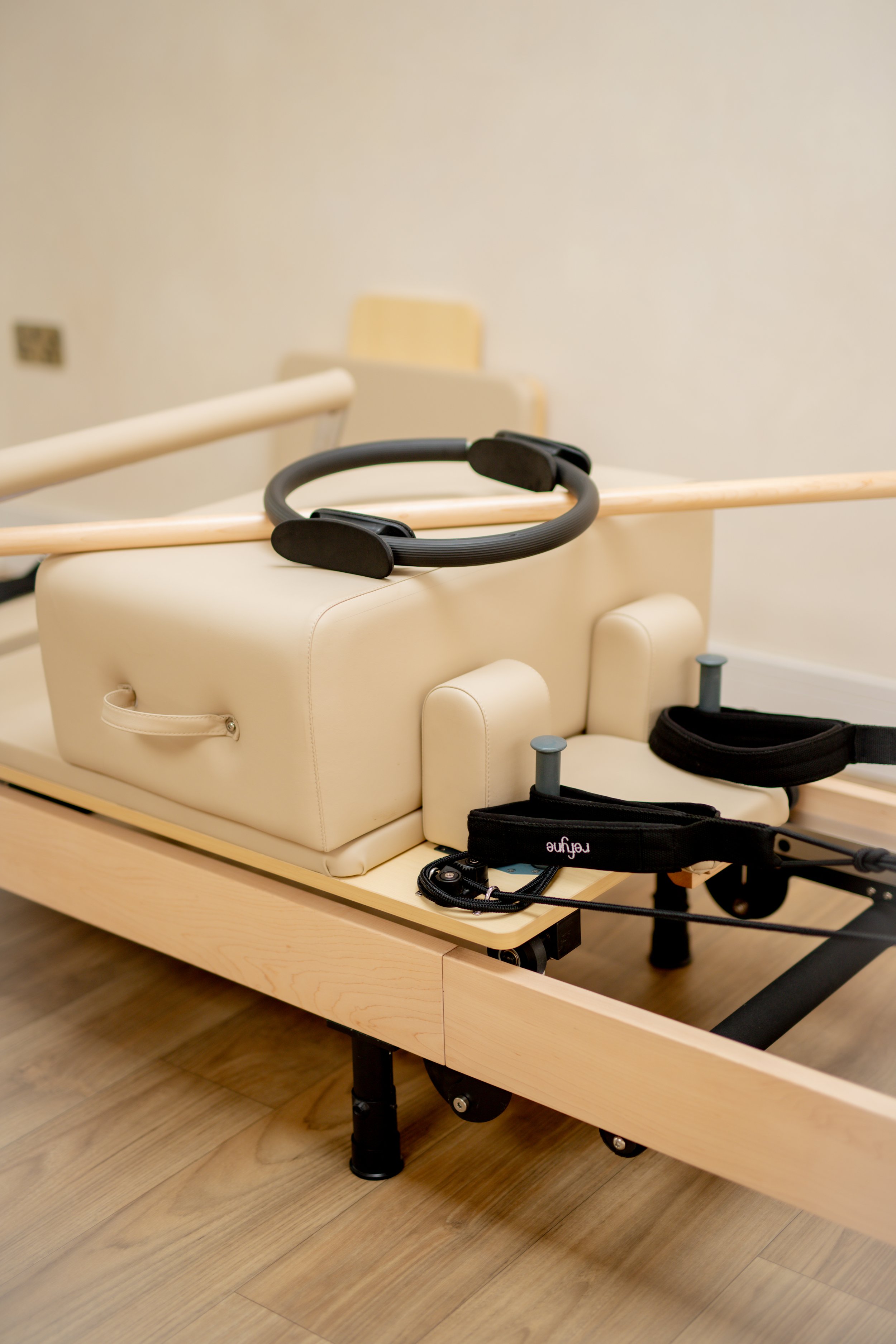 A specialized medical or therapy bed with attachments, including a padded cream-colored cushion, a black head strap, and a black cuff with a cable, situated on a wooden floor against a plain wall.