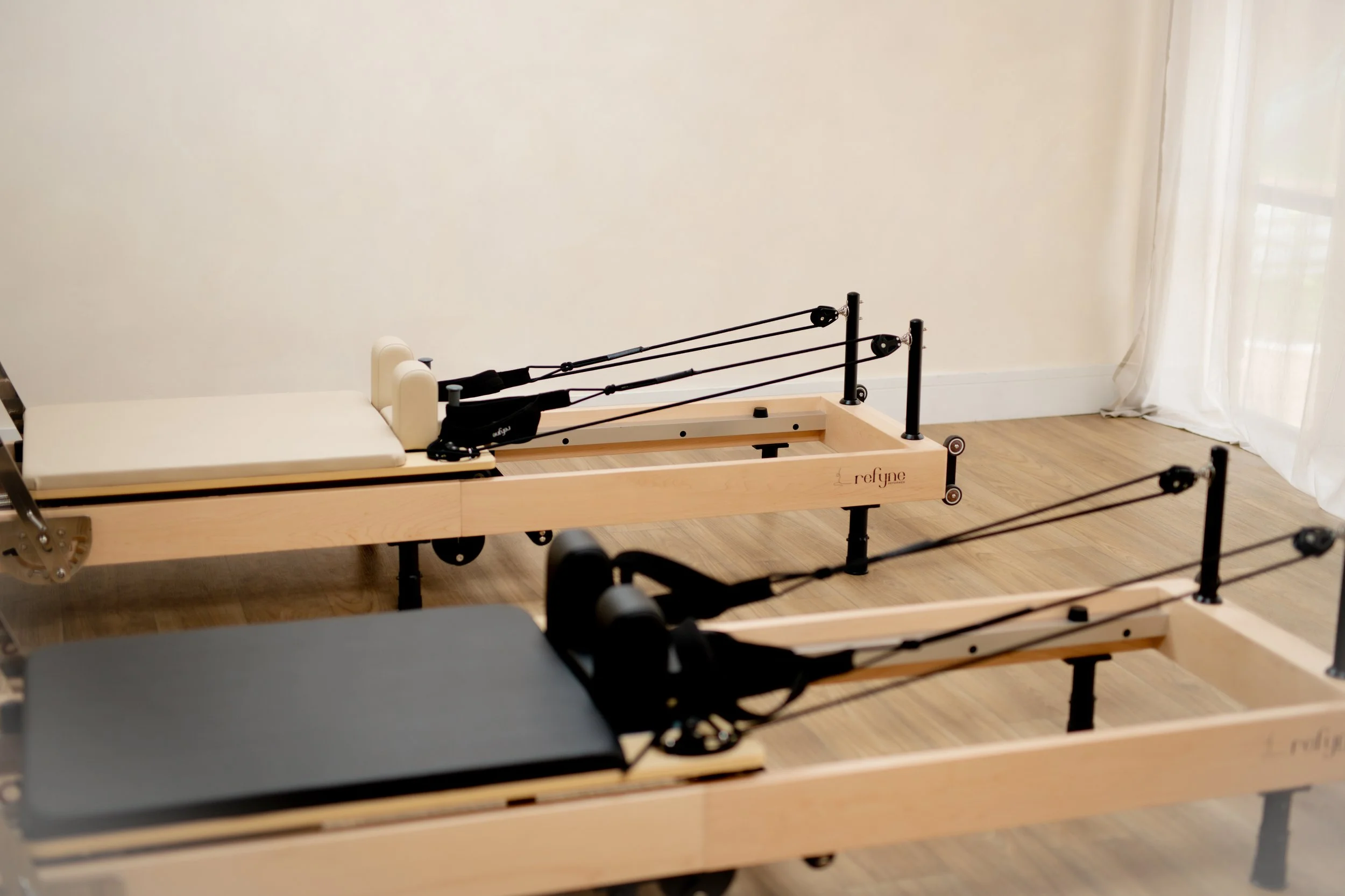 Two wooden reformer Pilates machines with black and white padded parts in a room with hardwood floors and white curtains.