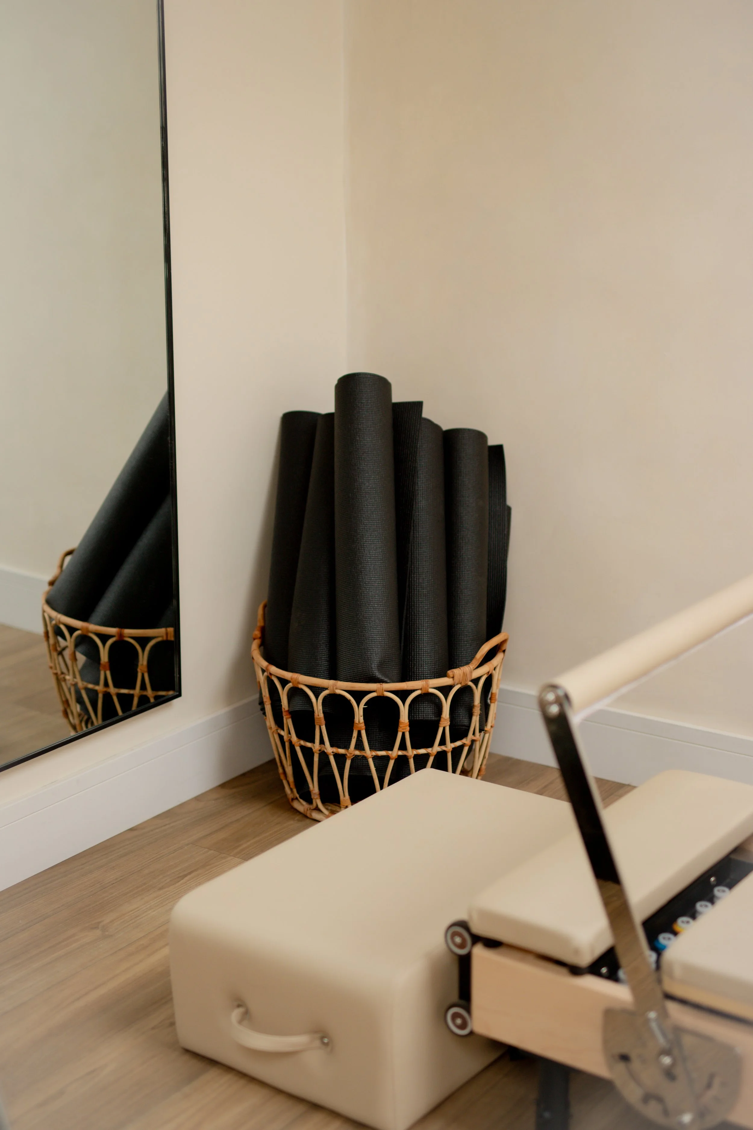 A corner of a room with a basket filled with rolled-up black yoga mats, a full-length mirror, and an adjustable massage or therapy table with a beige cushioned surface.