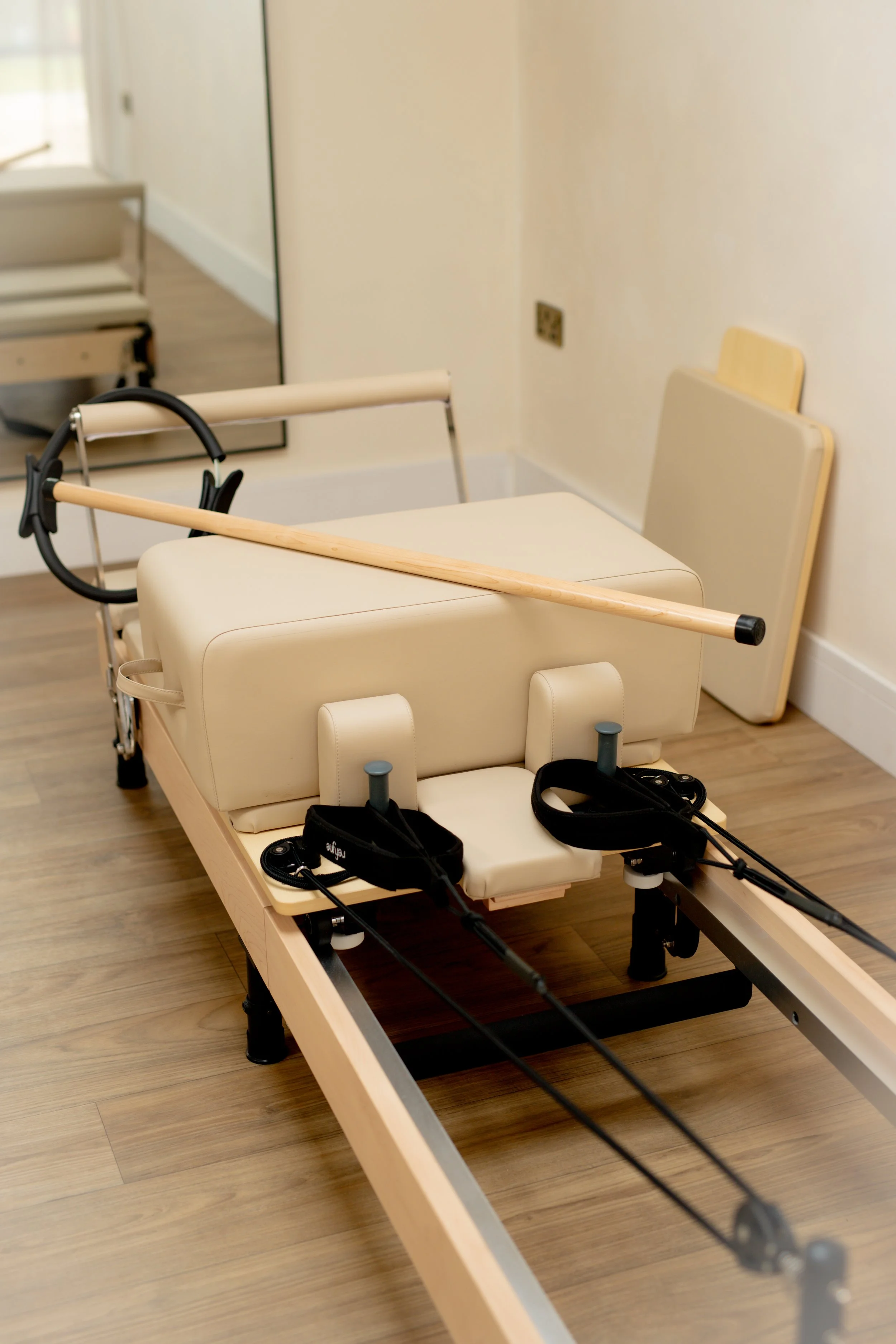 Hospital gurney with padded sides, a wooden staff resting on top, and medical restraints attached to the sides, located in a room with wooden floors and beige walls.