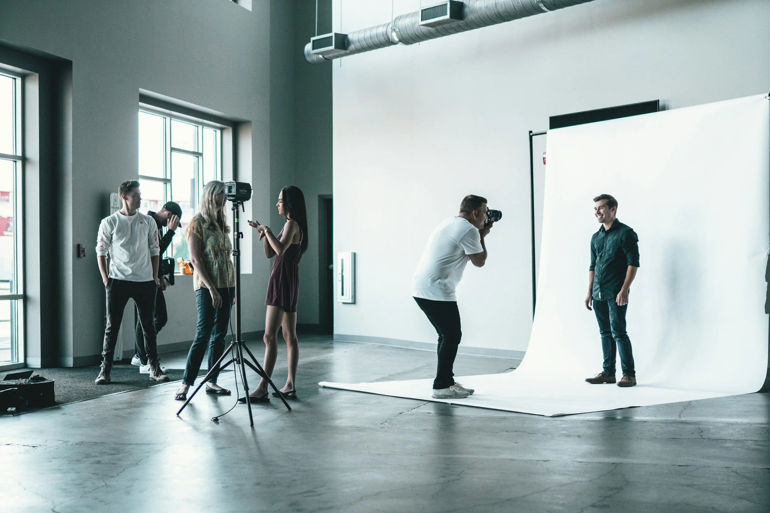 People doing a photoshoot in a studio with a white backdrop, a photographer taking pictures of a smiling man, and others preparing or adjusting the scene.