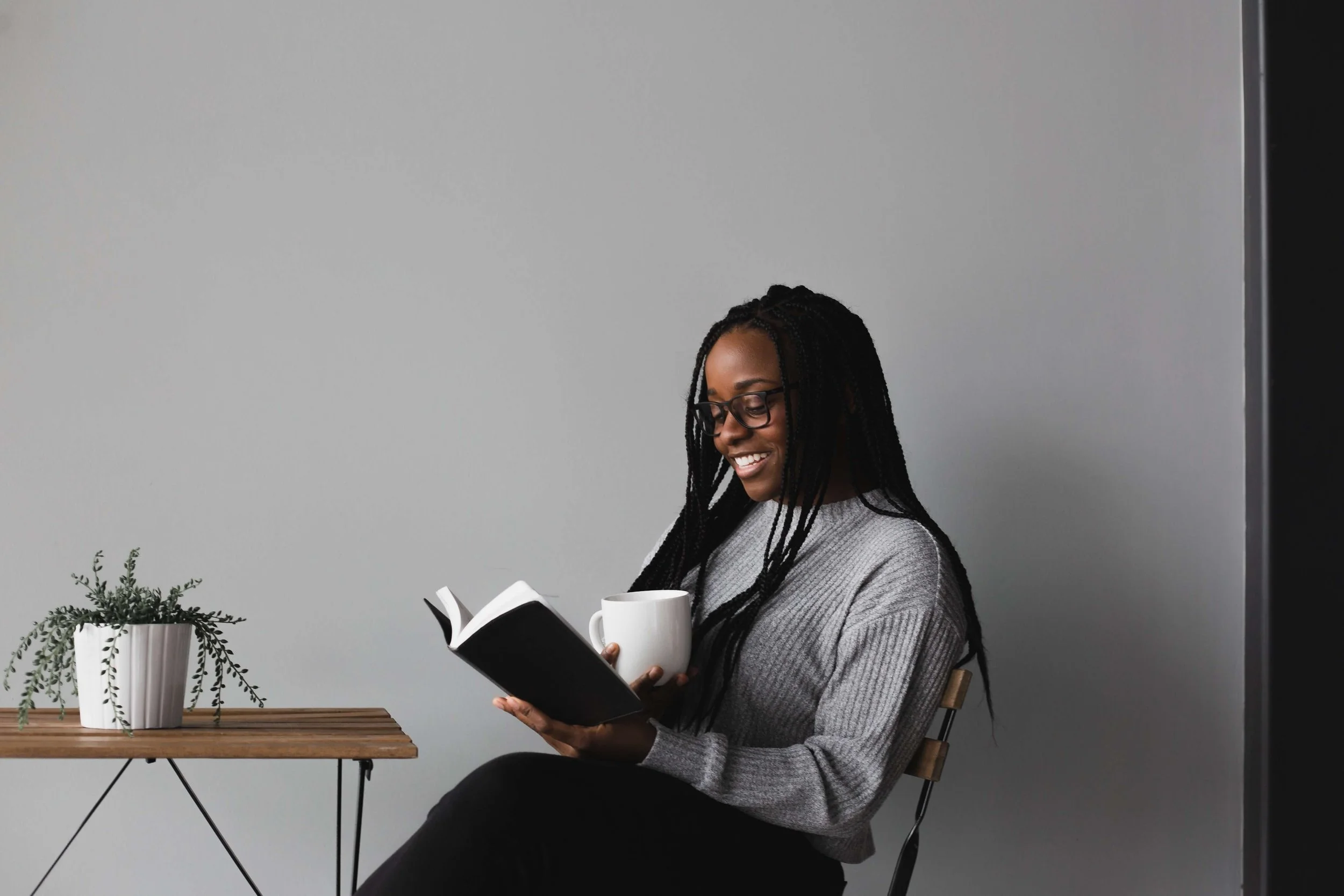 A woman with glasses and dreadlocks sitting on a chair, smiling while reading a book in her right hand and holding a white mug in her left hand, next to a small wooden table with a potted plant.