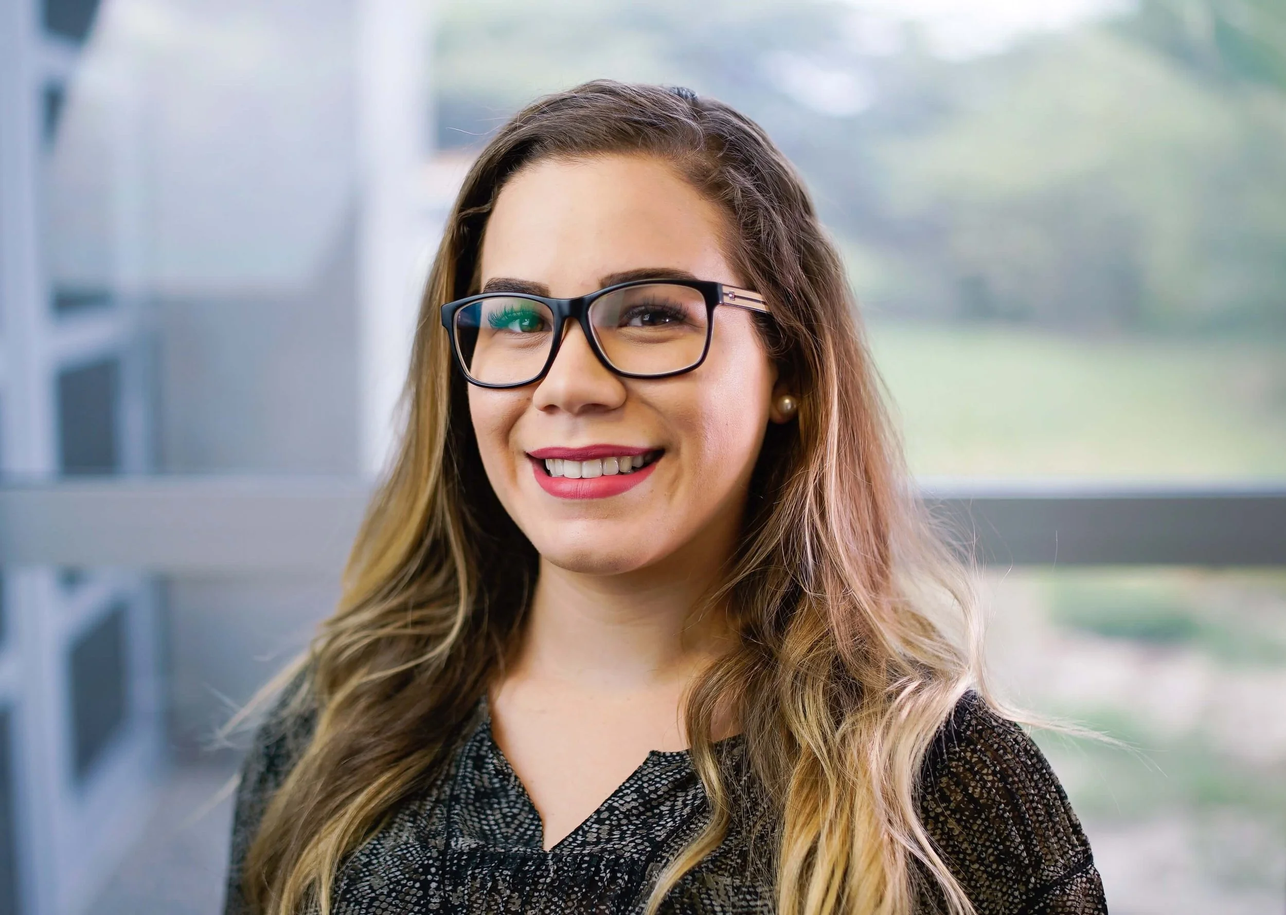A young woman with long hair, glasses, and lipstick smiling at the camera, standing indoors near a window with a blurred outdoor view in the background.