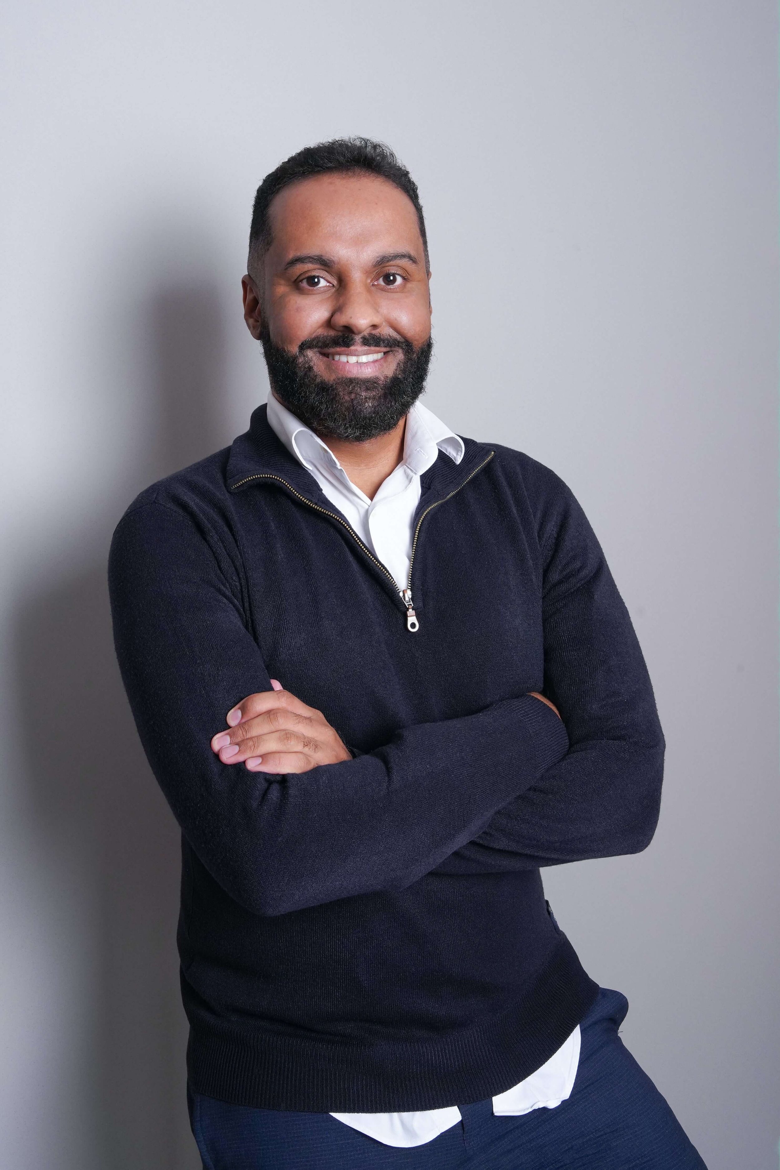 A smiling man with a beard and mustache standing against a plain gray wall, arms crossed, wearing a navy sweater with a zipper and white shirt underneath.