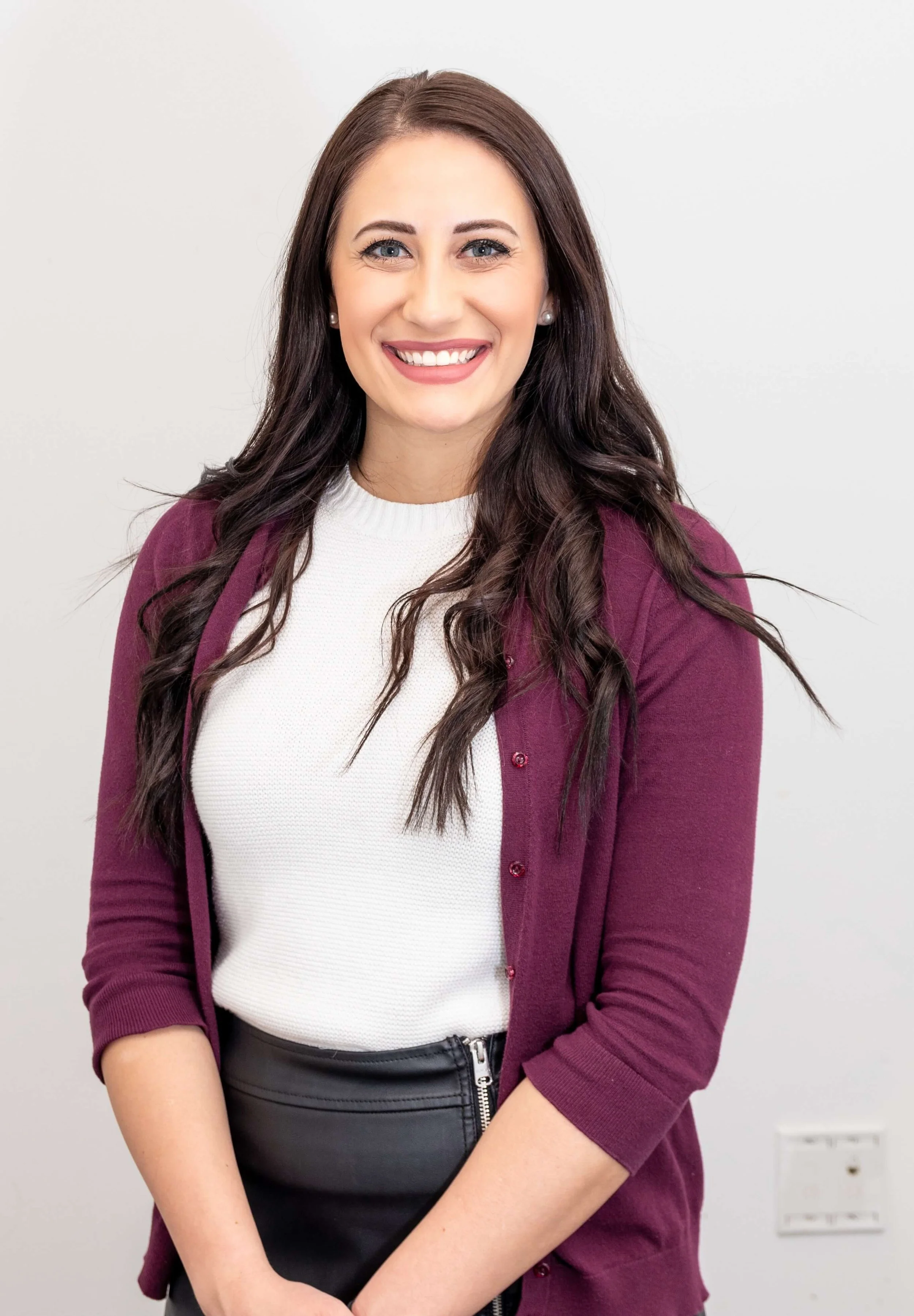 A young woman with long dark brown wavy hair, wearing a white top, burgundy cardigan, and black skirt, smiling against a plain light background.