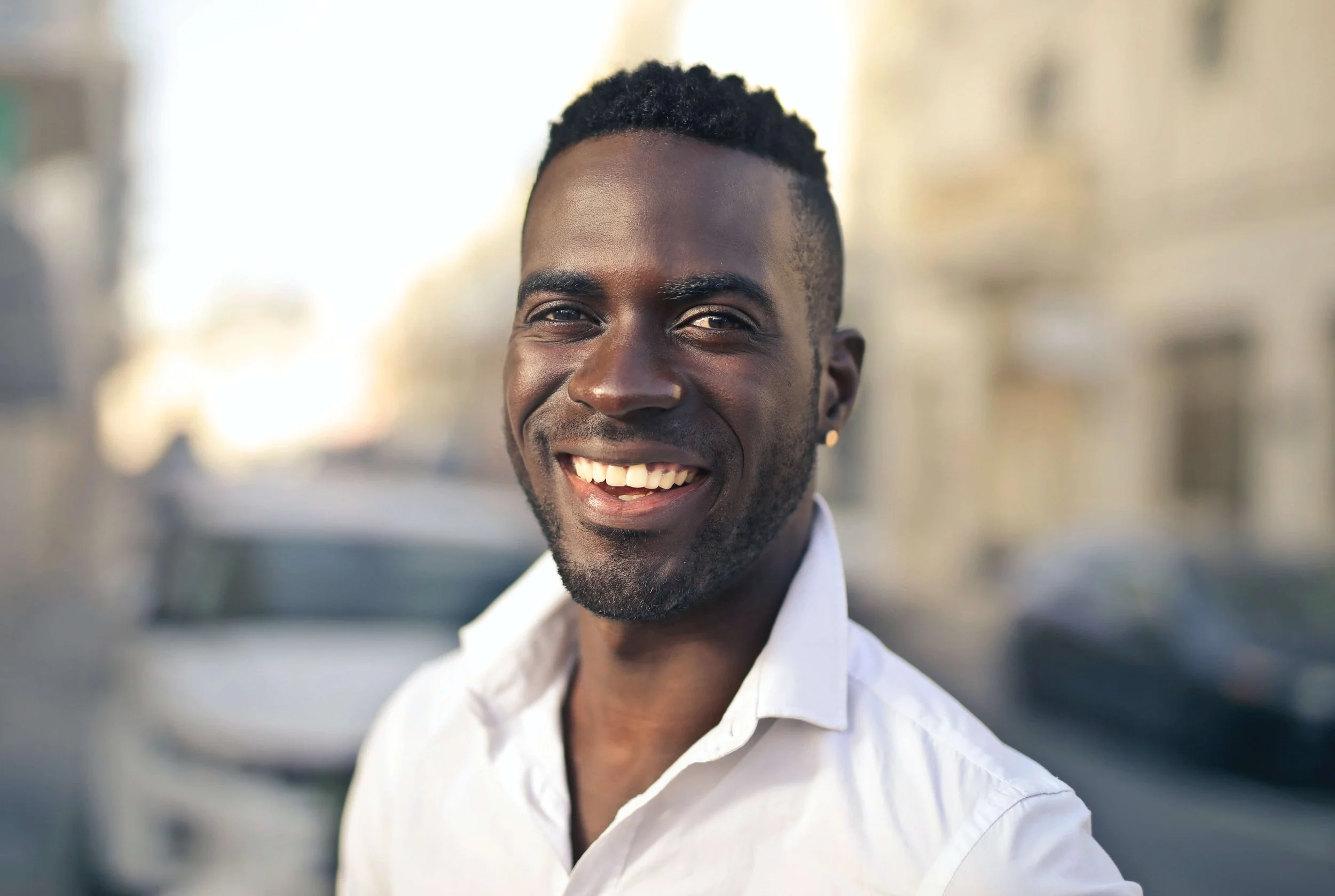 A smiling man with dark curly hair, wearing a white shirt, standing outdoors on a sunny day with blurred buildings and vehicles in the background.