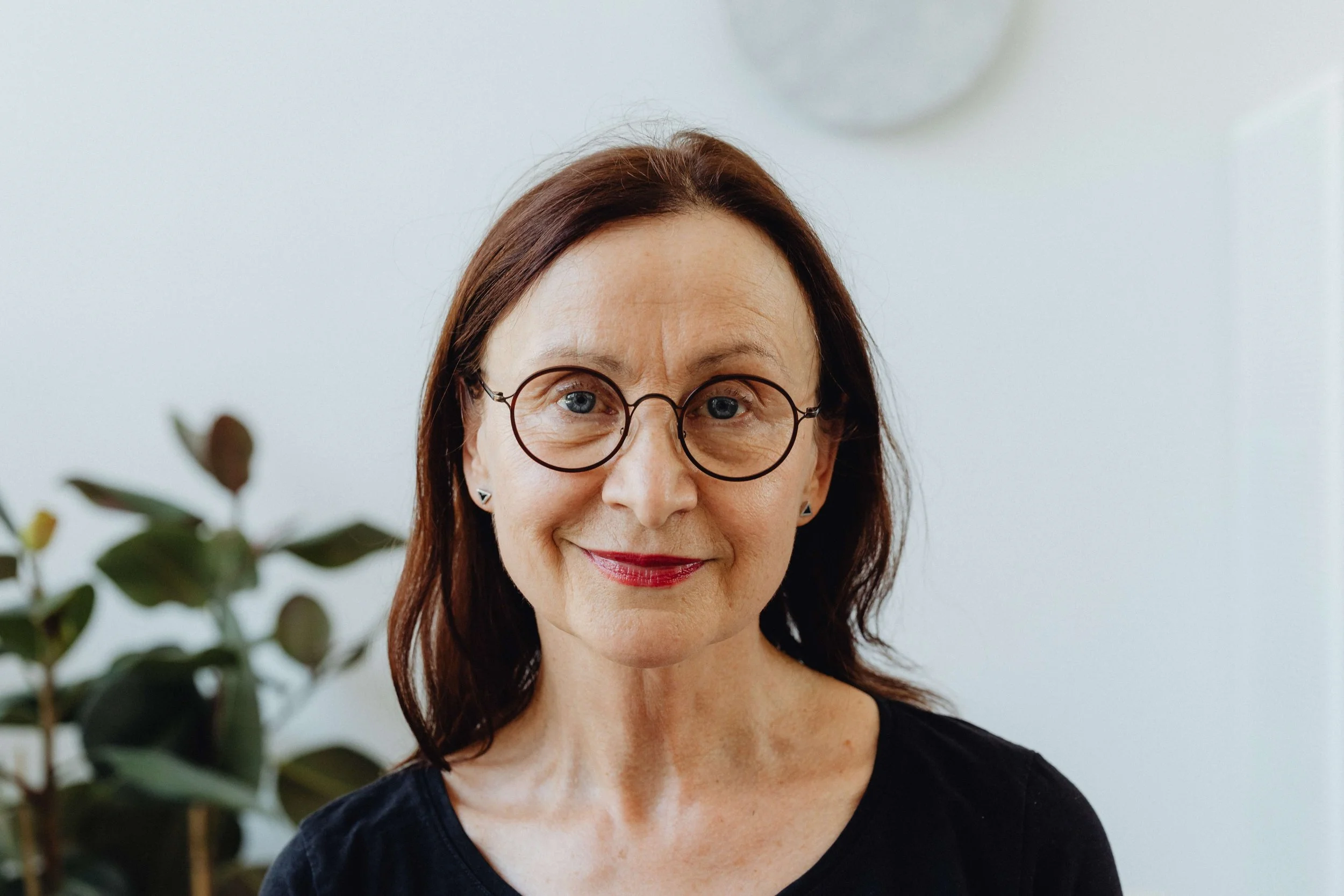 A middle-aged woman with shoulder-length brown hair, wearing glasses and a black top, smiling indoors with a plant and a white wall in the background.