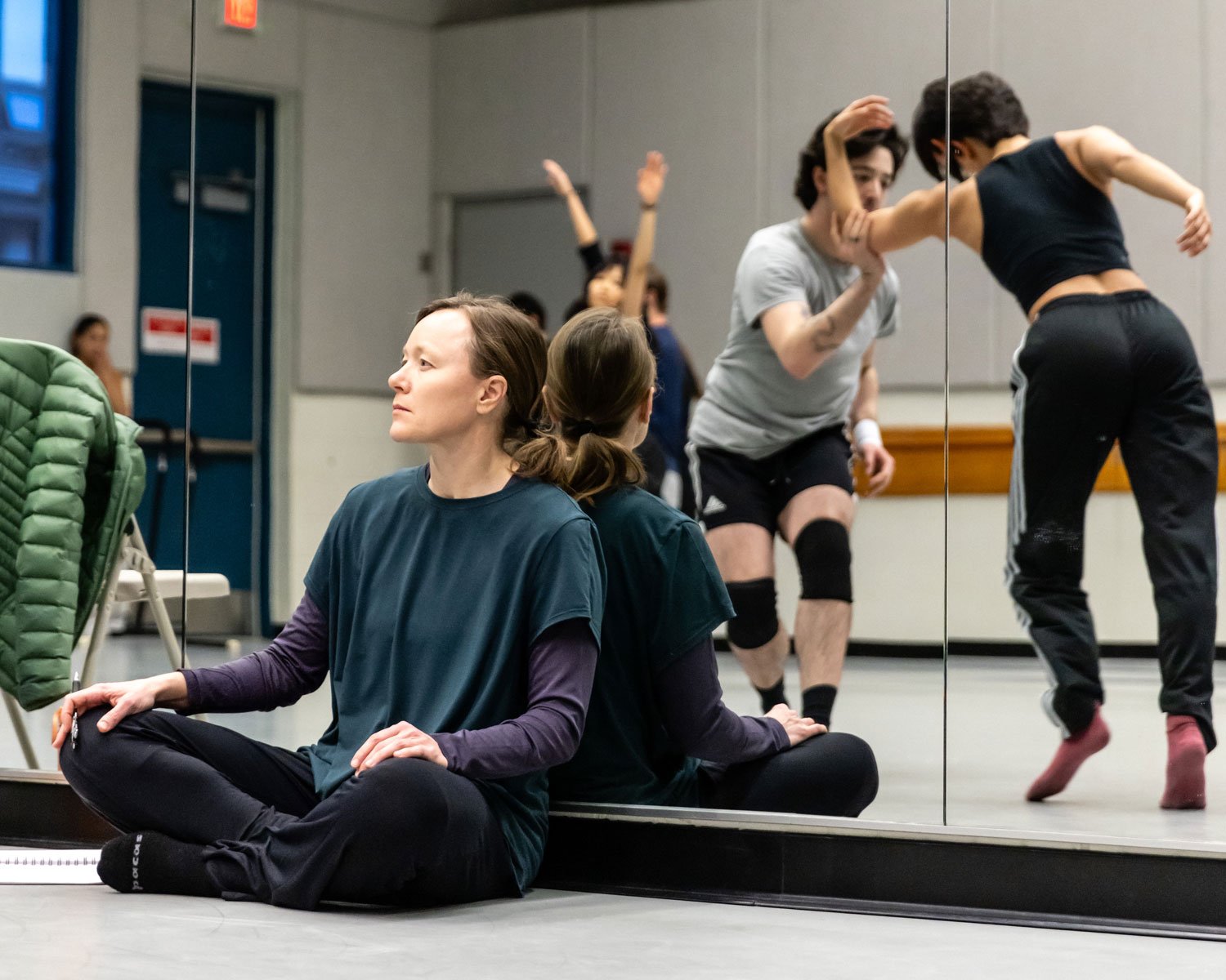 A woman sitting cross-legged on the floor in front of a mirror, practising meditation or yoga. In the background, two women are dancing or wrestling near the mirror.