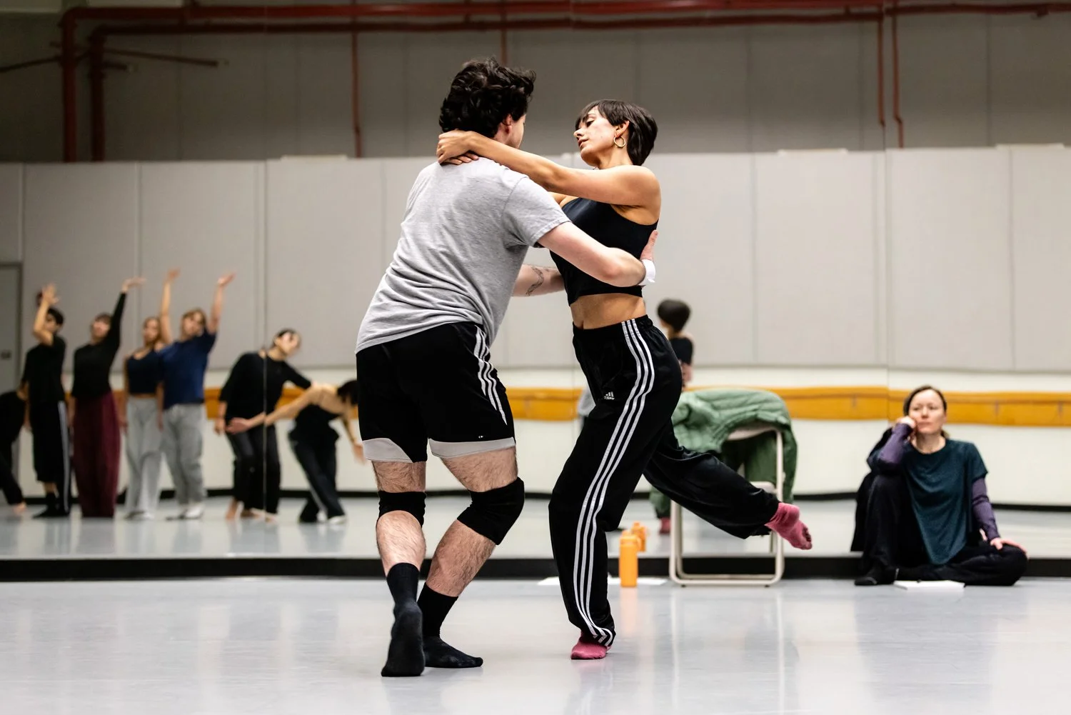 Two people are dancing closely in the foreground while a group of people in the background are stretching or practicing dance moves in a studio.