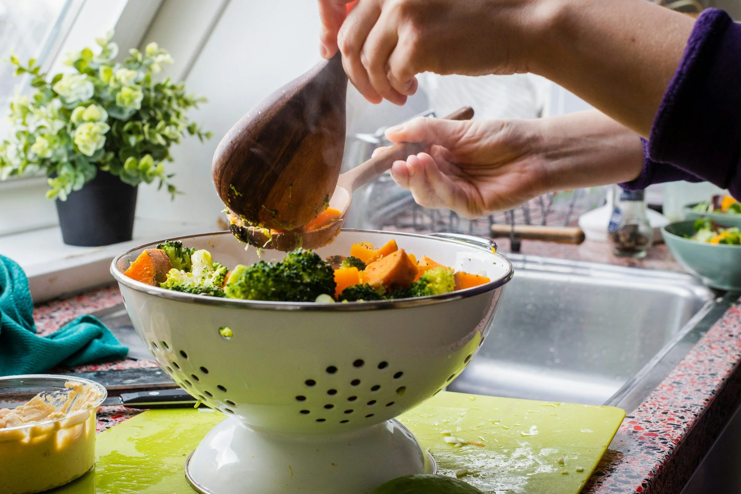 Hands pouring steamed vegetables into a white colander in a kitchen