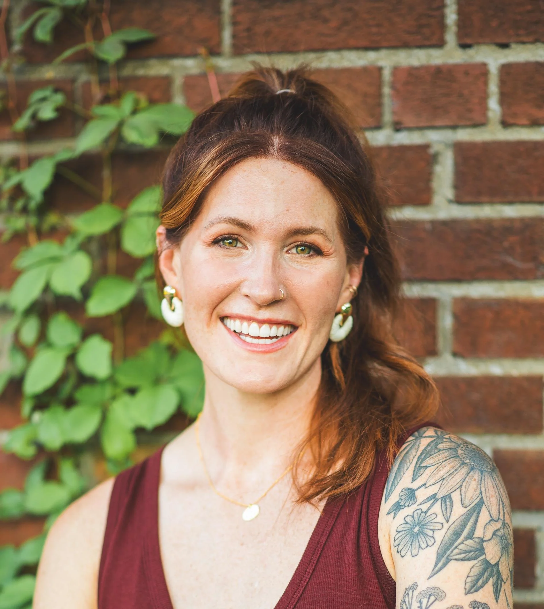 Close-up of a smiling woman with green eyes, brown hair, a nose piercing, and a tattoo sleeve on her left arm, standing against a brick wall with green leafy plants.
