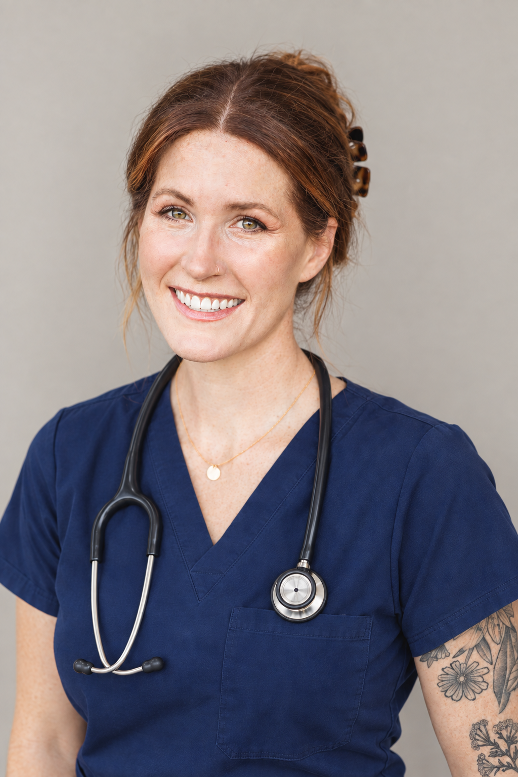A smiling female healthcare professional with red hair, wearing navy scrubs and a stethoscope around her neck, standing against a plain gray background.