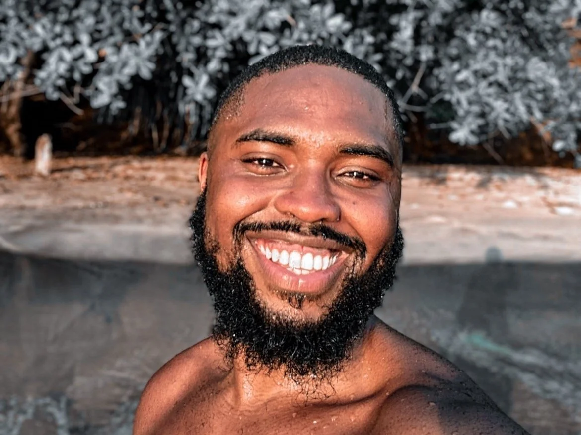 Homem sorridente com barba em um ambiente ao ar livre.