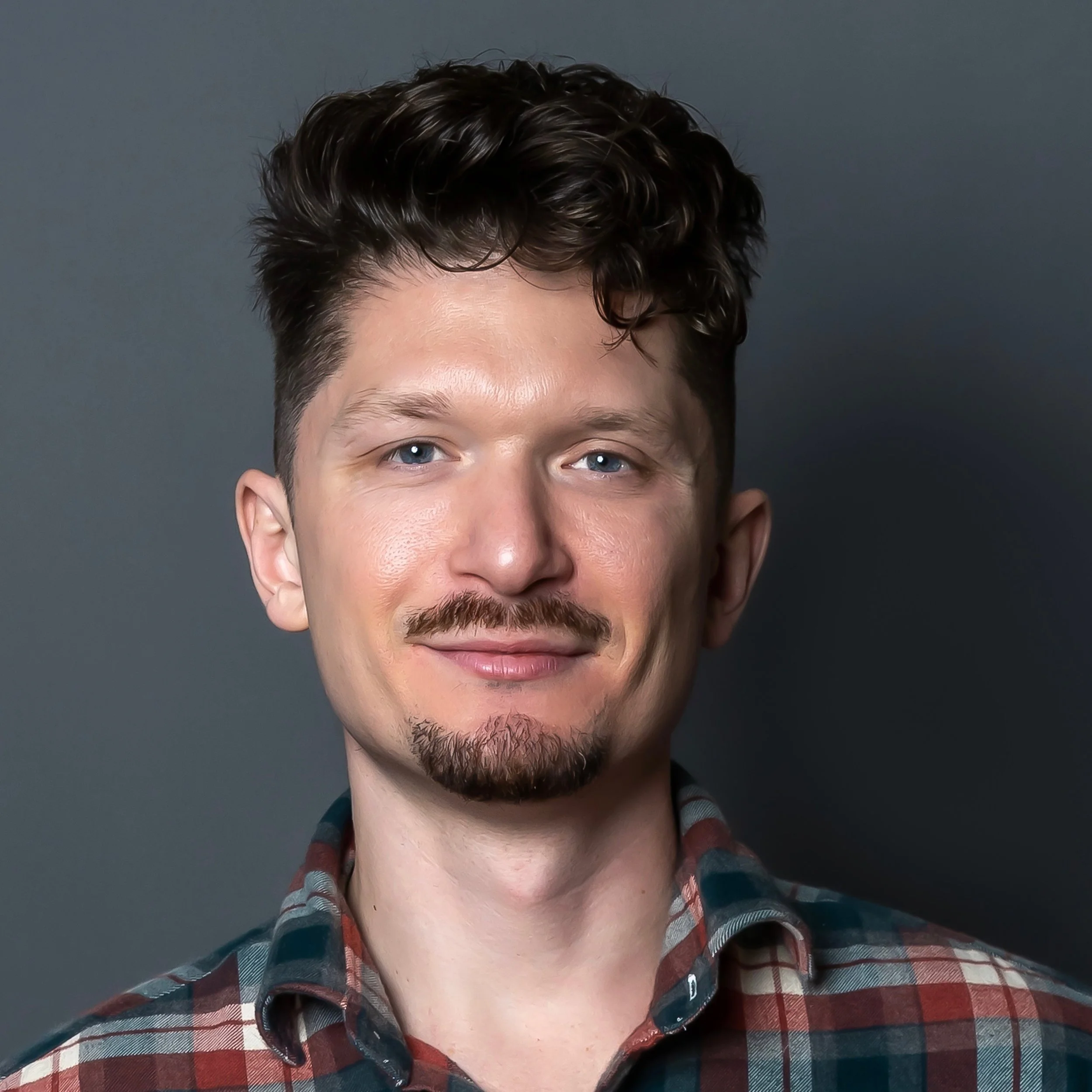 A young man with curly dark hair, blue eyes, and a trimmed beard, wearing a plaid shirt, smiling at the camera against a dark gray background.
