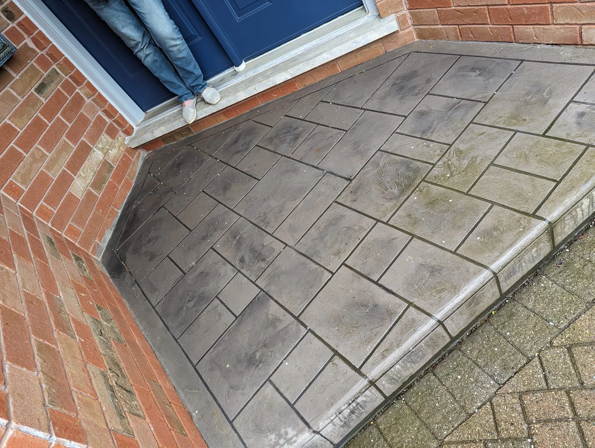 Close-up of a house entrance with a concrete step, patterned with rectangular stones, surrounded by brick and paver surfaces, with a person standing near the door.