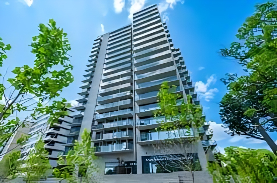 A tall modern apartment building viewed from the ground up with green trees in the foreground and a partly cloudy blue sky.