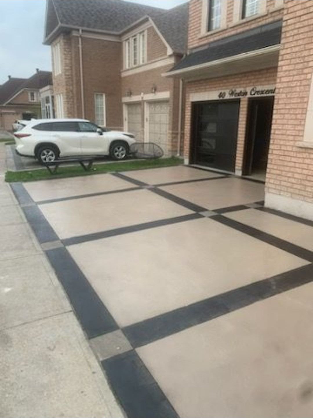 Driveway with a geometric pattern of large beige tiles bordered by black lines, leading to a garage and an entrance to a brick house.