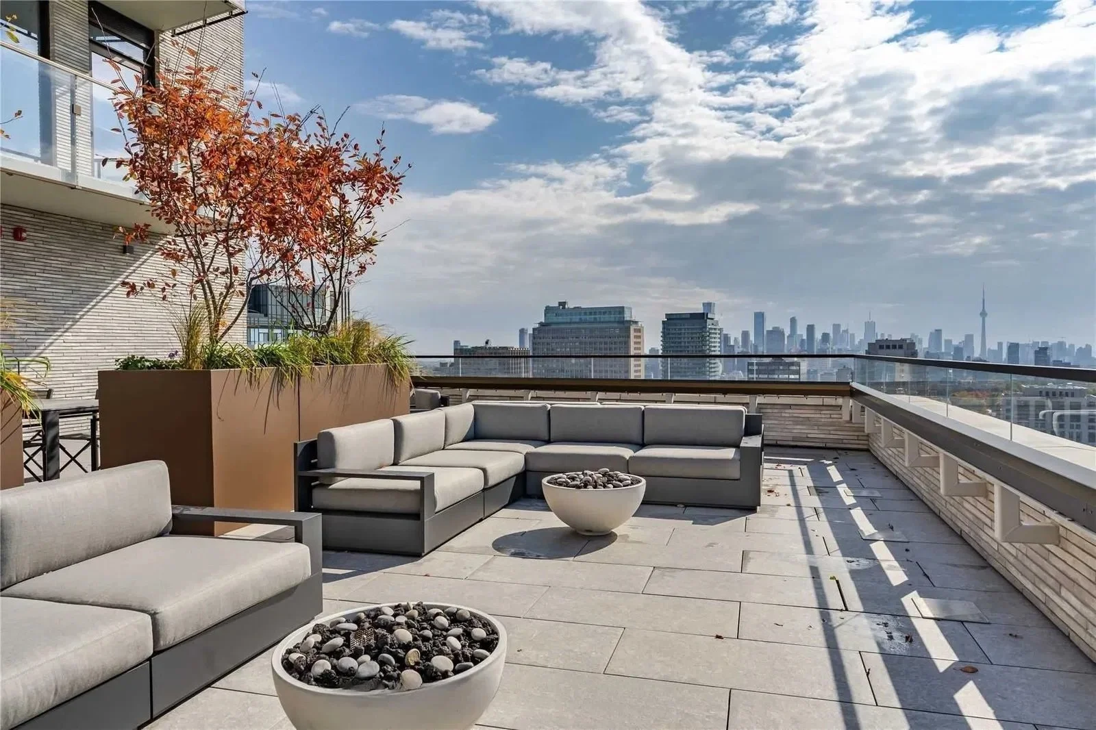 Rooftop patio with modern gray outdoor sofas, potted plants, and a city skyline view with tall buildings and a partly cloudy sky.