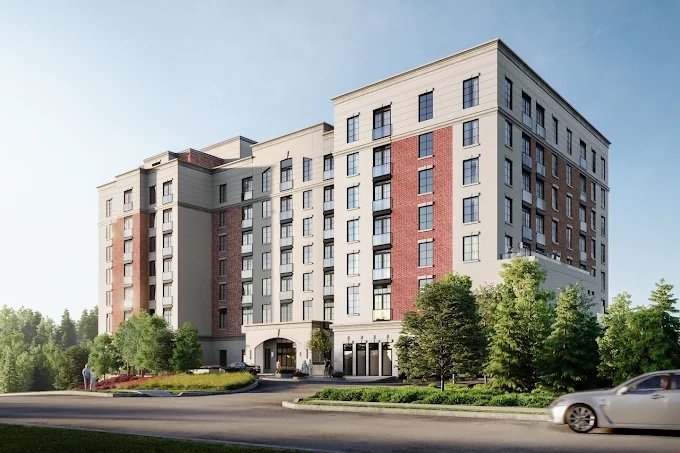 A multi-story residential building with beige and red brick exterior, surrounded by green trees and a small landscaped area in front, with a car driving on the street.