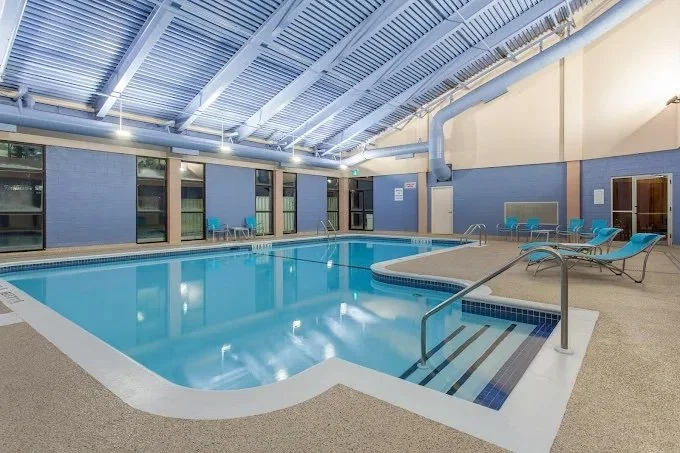 Indoor swimming pool with blue walls, lounge chairs, and large windows, under a metal ceiling with lighting.