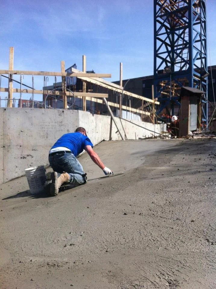 A construction worker wearing a blue shirt and jeans is smoothing wet concrete on the ground at a construction site, with wooden scaffolding and a metal tower in the background under a clear blue sky.