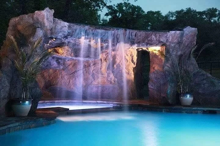A backyard pool with a faux rock waterfall feature illuminated at dusk, with water flowing over the rocks into the pool.