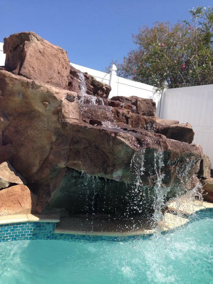 Swimming pool with a rock waterfall feature with water cascading down rocks into the pool, surrounded by a white fence and a tree with pink flowers, under a clear blue sky.
