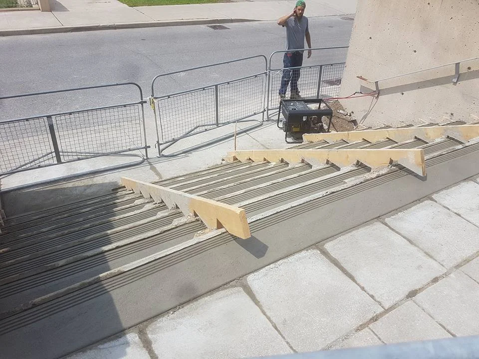 A construction site with a set of stairs under renovation, a worker standing in the background, and equipment including a saw placed on a temporary barrier.