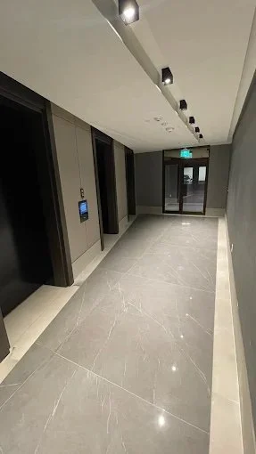 Empty modern elevator lobby with gray walls, large floor tiles, and black ceiling lights, featuring elevator doors and an emergency exit door at the end.