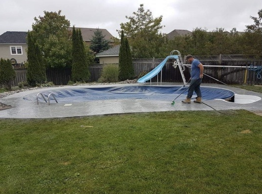 Person cleaning an empty in-ground backyard swimming pool on a cloudy day, with houses and trees in the background.