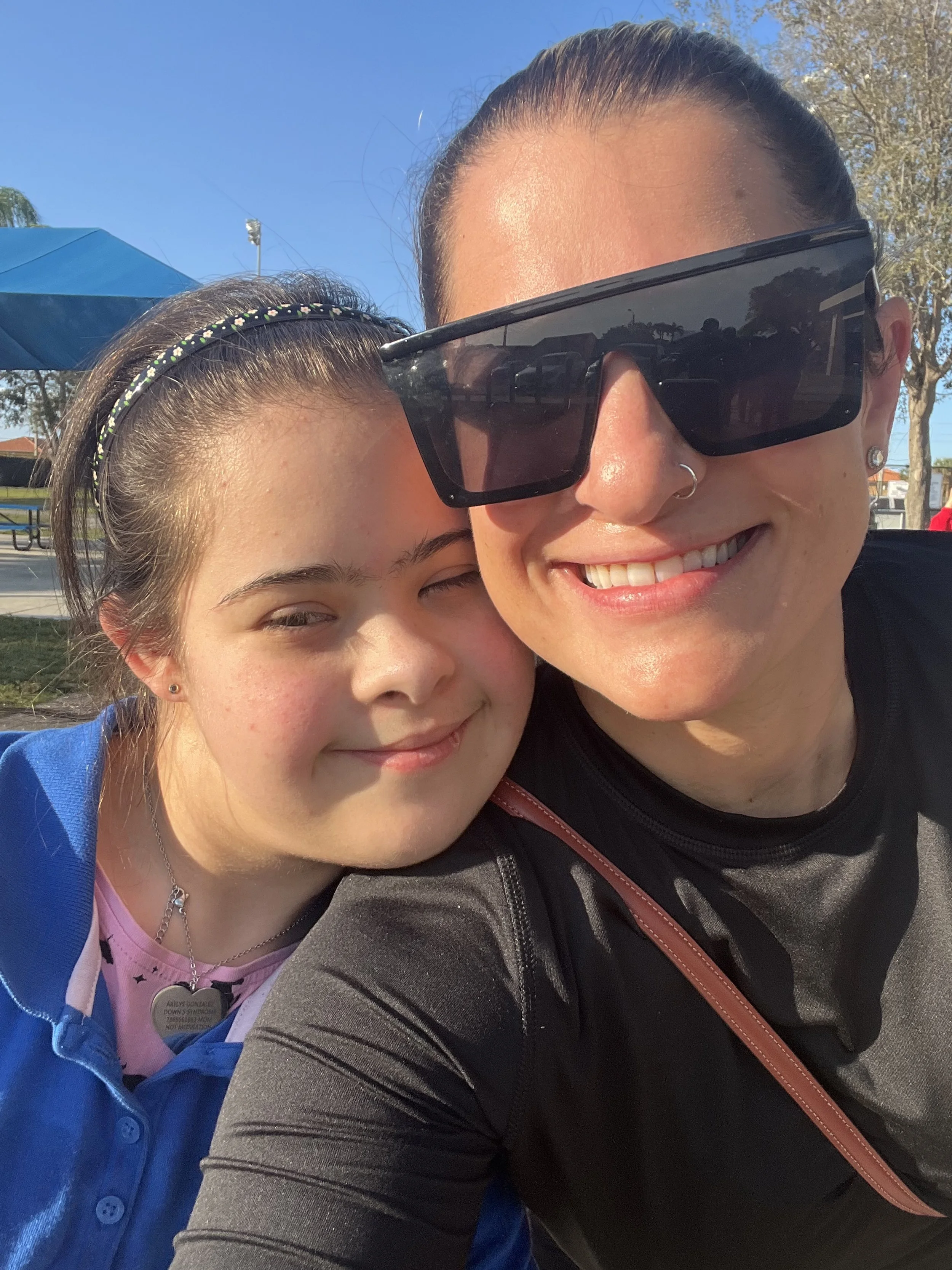 A woman and a girl taking a close-up selfie outdoors. The woman is wearing large sunglasses, a nose ring, and a black shirt, smiling. The girl is smiling with her eyes closed, wearing a blue jacket, a necklace, and a headband.