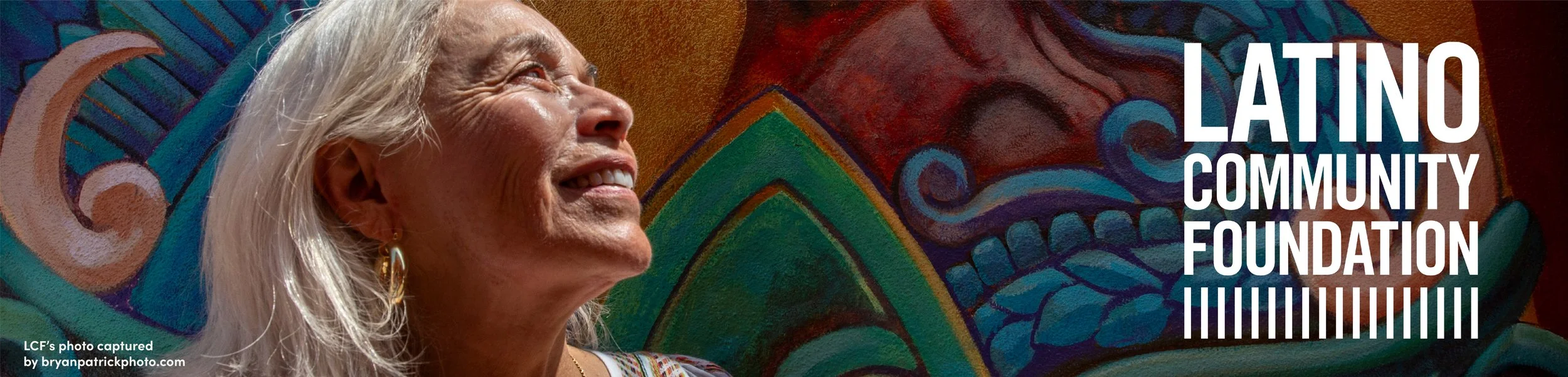 An elderly woman with long gray hair smiling and looking up, in front of a colorful mural background with the text 'Latino Community Foundation' and a barcode.
