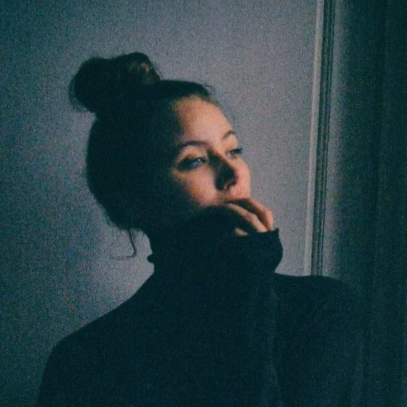 A woman with her hair in a bun, wearing a black turtleneck, partially covering her mouth with her hand, standing against a gray wall.