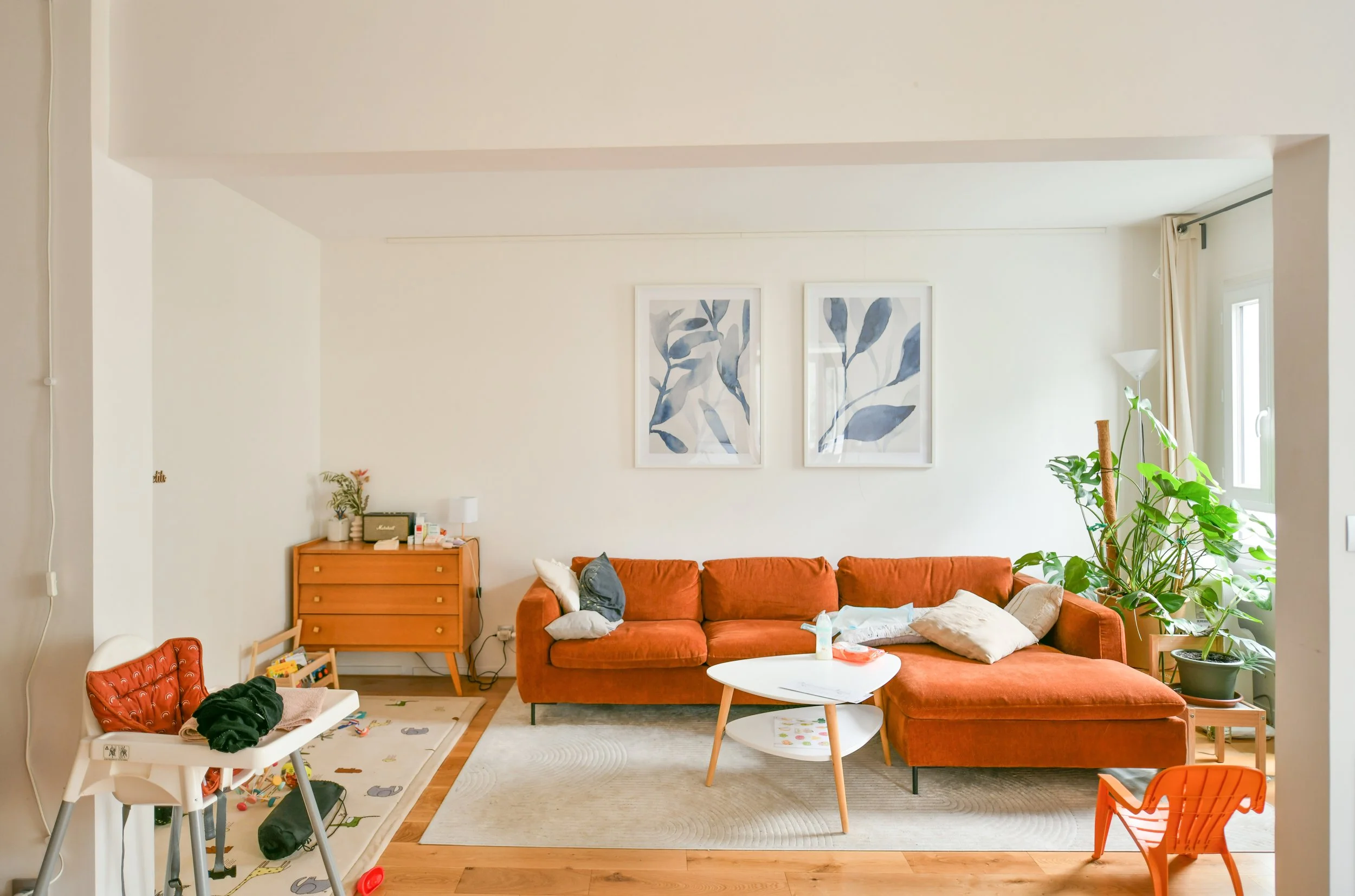 Séjour avec canapé orange, tableau botanical sur le mur, plantes vertes près de la fenêtre, table basse blanche, tapis beige, meubles en bois, décor simple et lumineux.