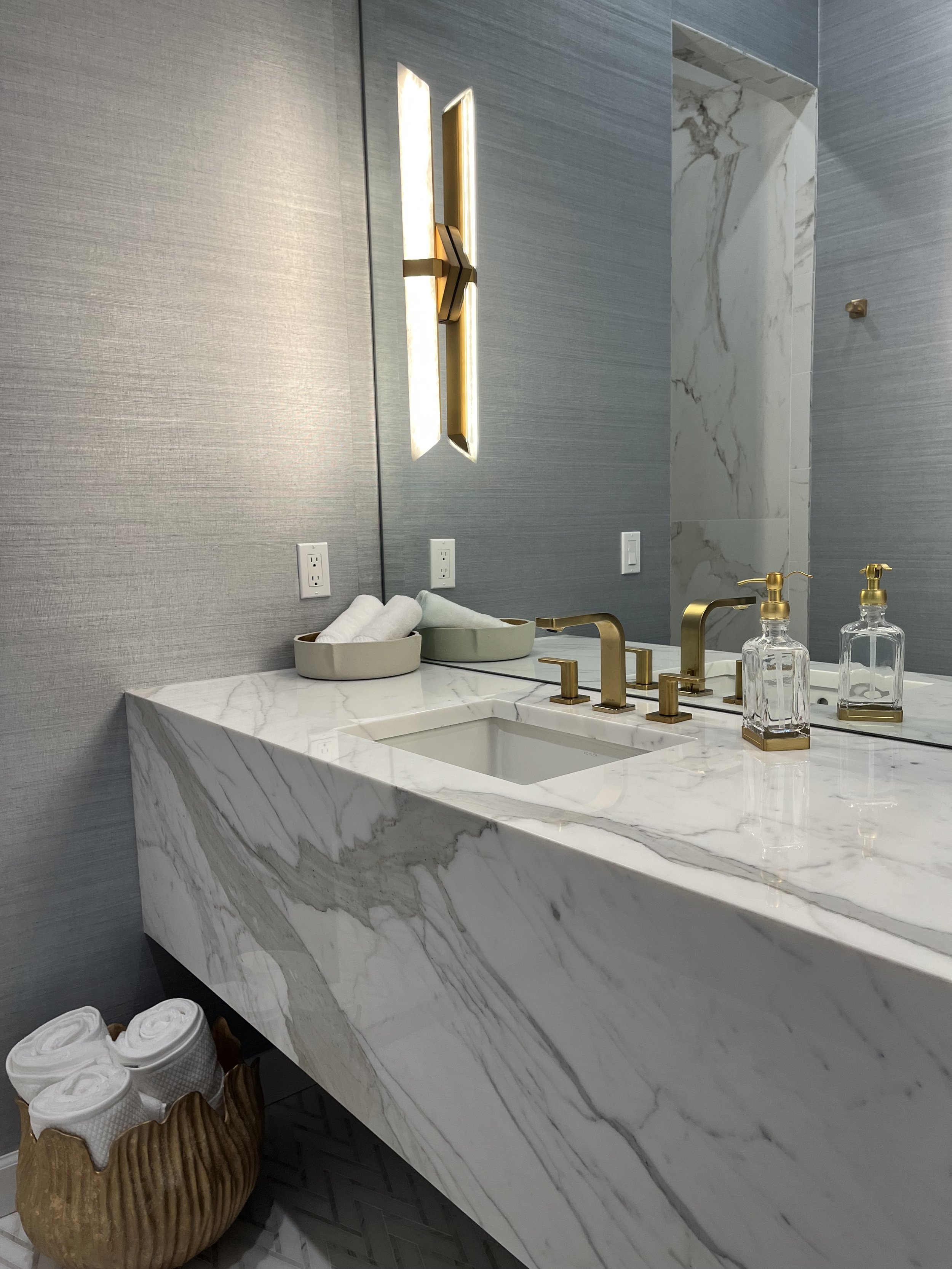 Modern bathroom with a marble sink, gold fixtures, and wall mirror. Decorative lighting and folded towels in a basket are present. The wall has a textured finish.