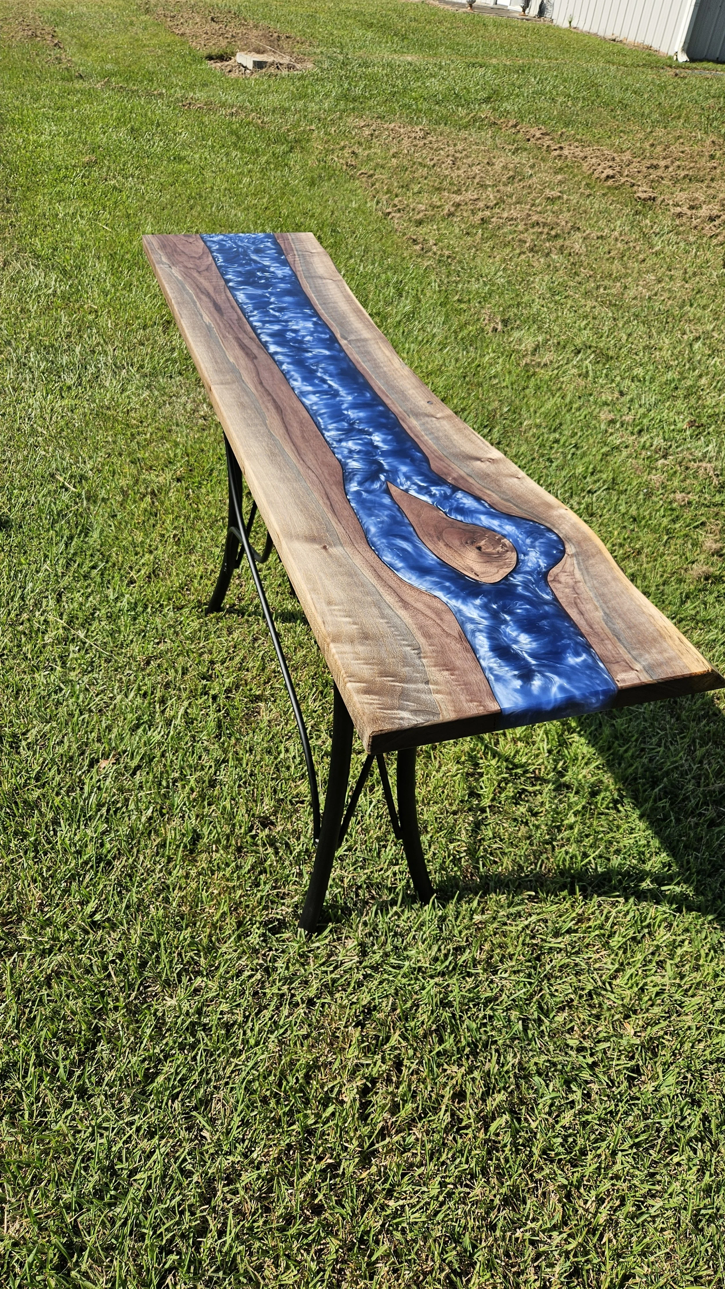 Wooden table with a resin river design, set on grass outdoors, with a building in the background.