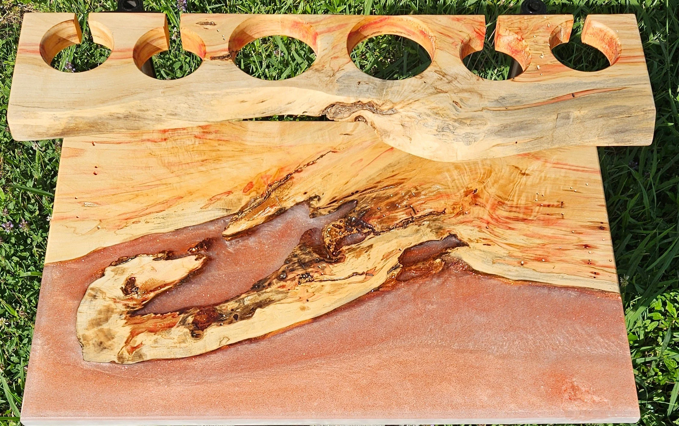 A wooden and red epoxy wine serving board resting on grass, with a wooden chess piece rack on top, designed with circular holes to hold chess pieces.