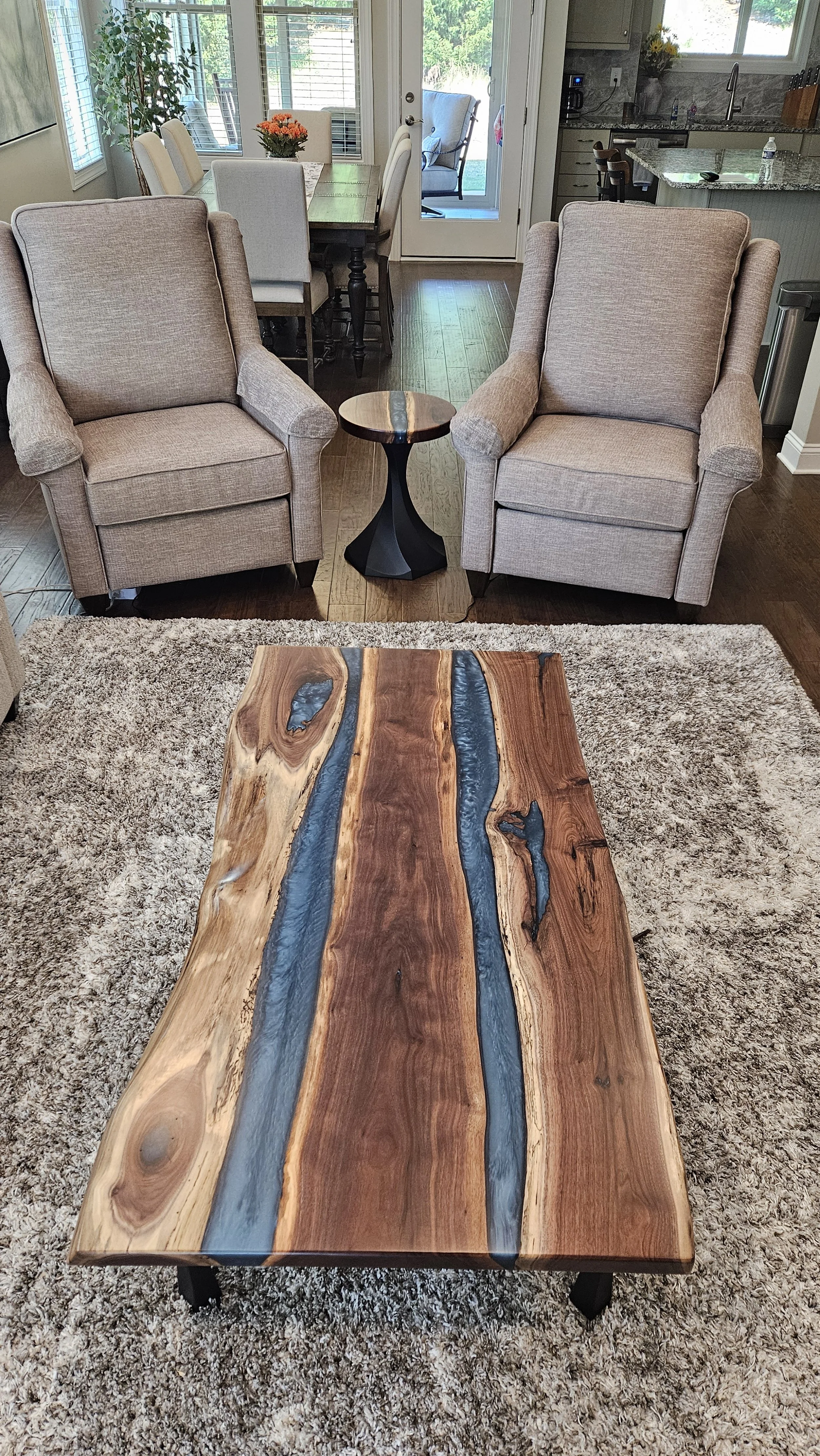 Living room with a rustic wood and epoxy coffee table, two beige armchairs, a small round side table, a dining table with chairs, and a view of the kitchen in the background.