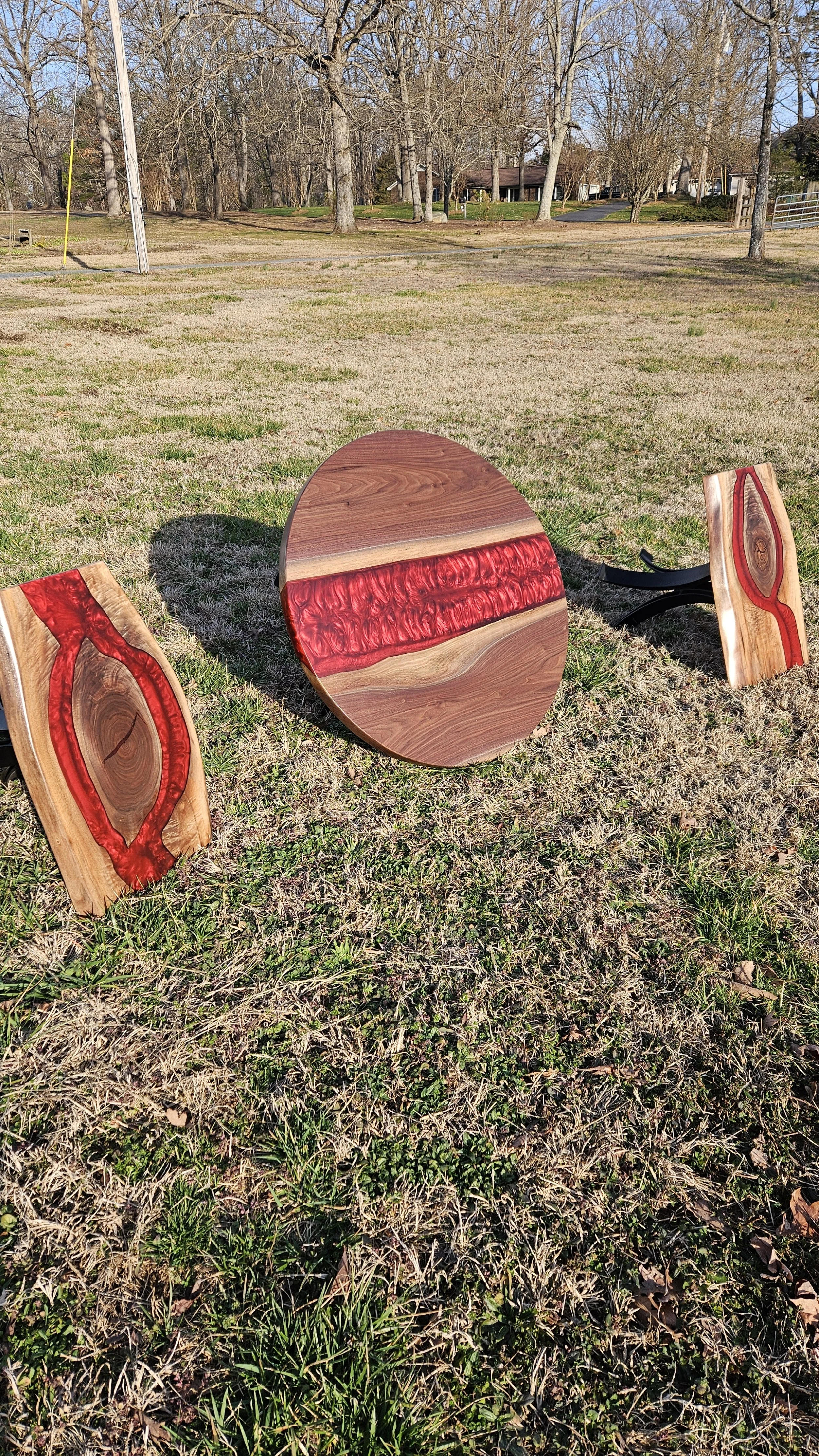 Three pieces of wooden furniture with red resin inlays on a grassy field, trees and houses in the background.