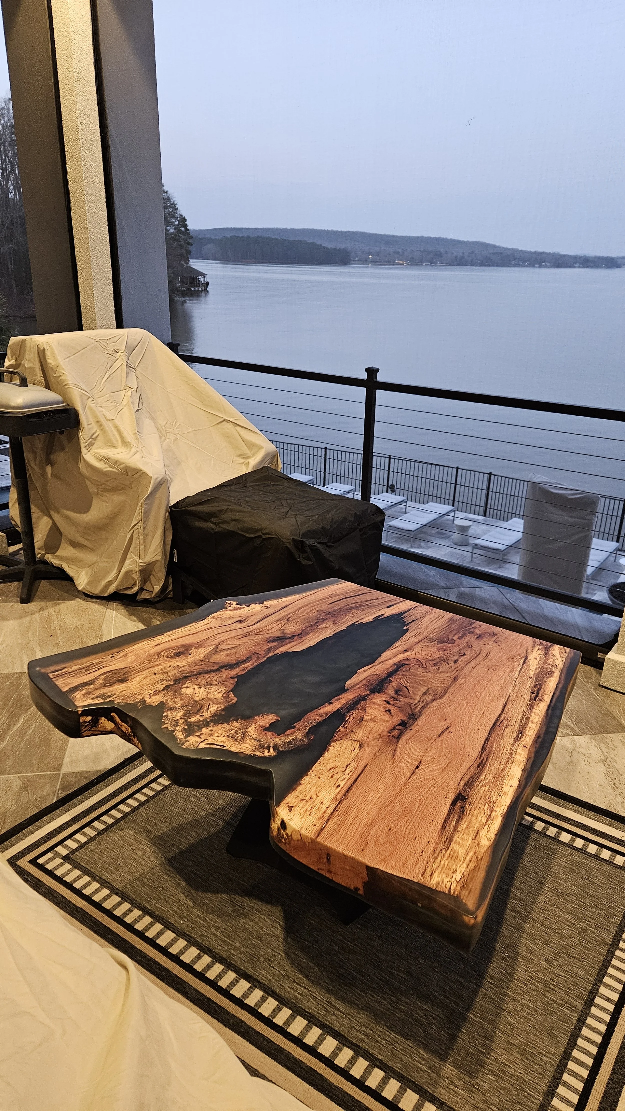 A wooden and resin coffee table on a rug with a lakeside view in the background.