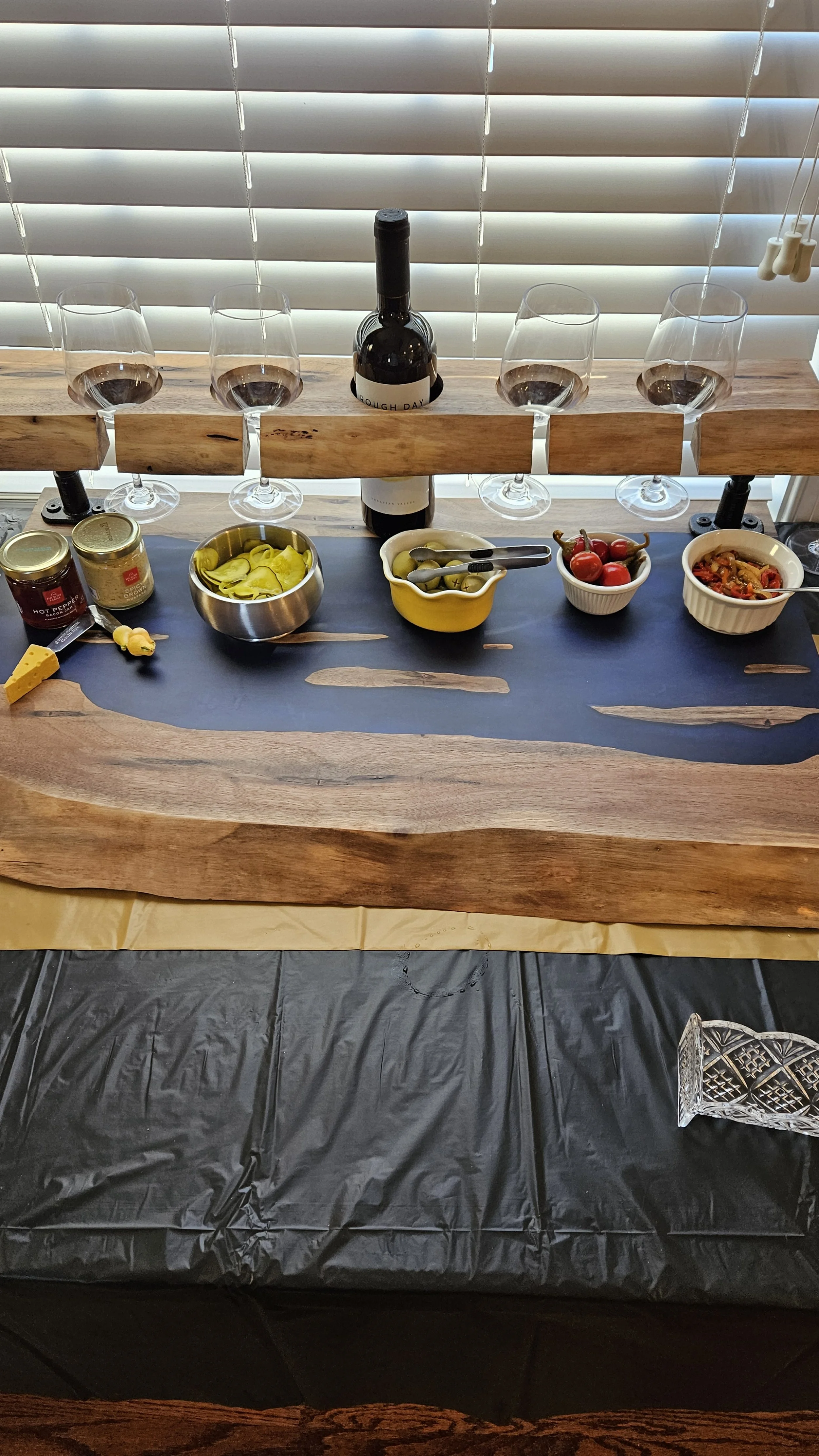 A gourmet wine and cheese tasting setup with five glasses of red wine, bowls of pickles, grapes, and spiced nuts, condiments, and a bottle of wine on a wooden and black surface with a striped window blind background.
