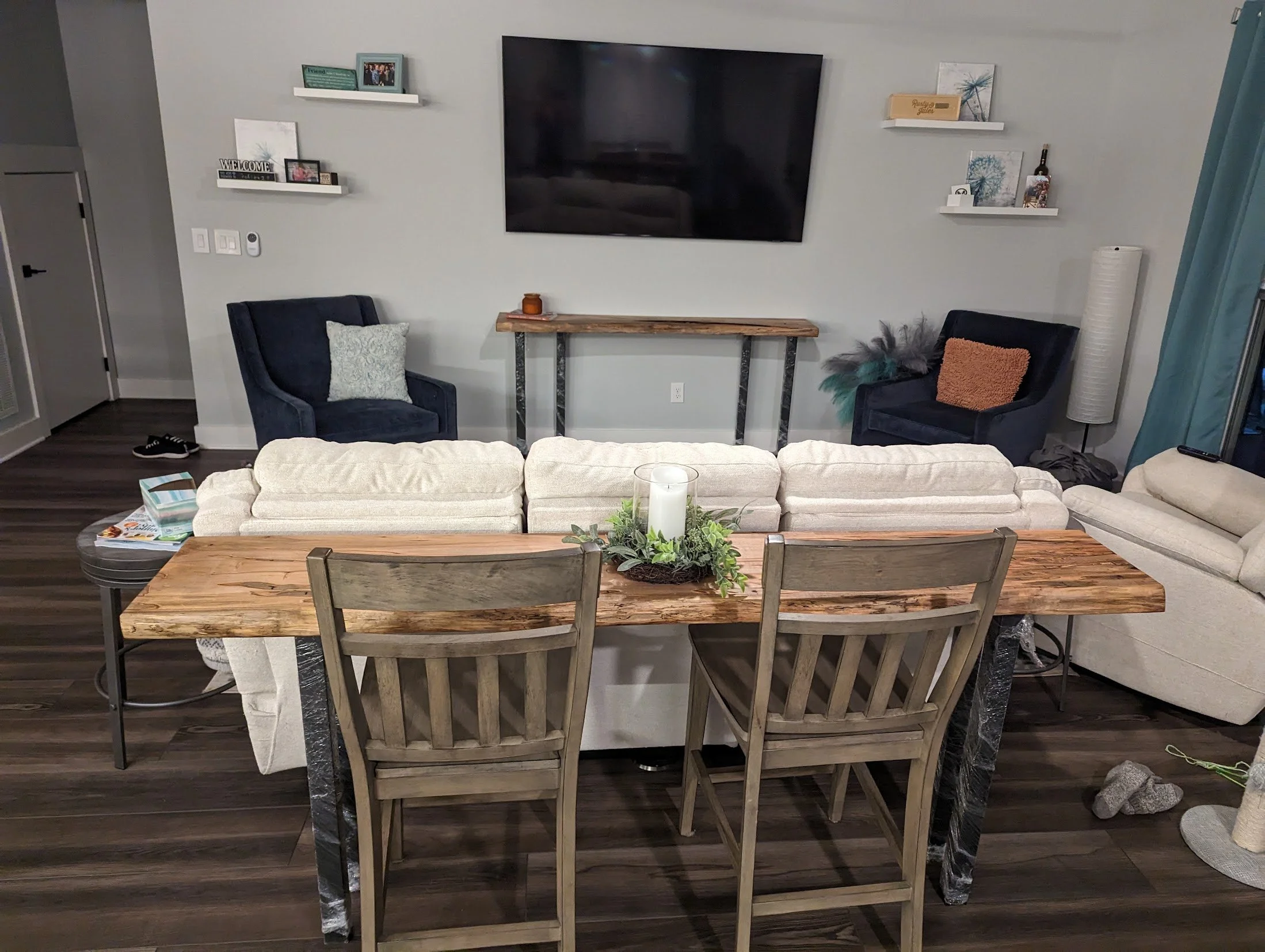 Living room with a wooden live edge dining table, beige sofa, two navy blue armchairs, wall-mounted TV, and decorative shelves on a light gray wall.