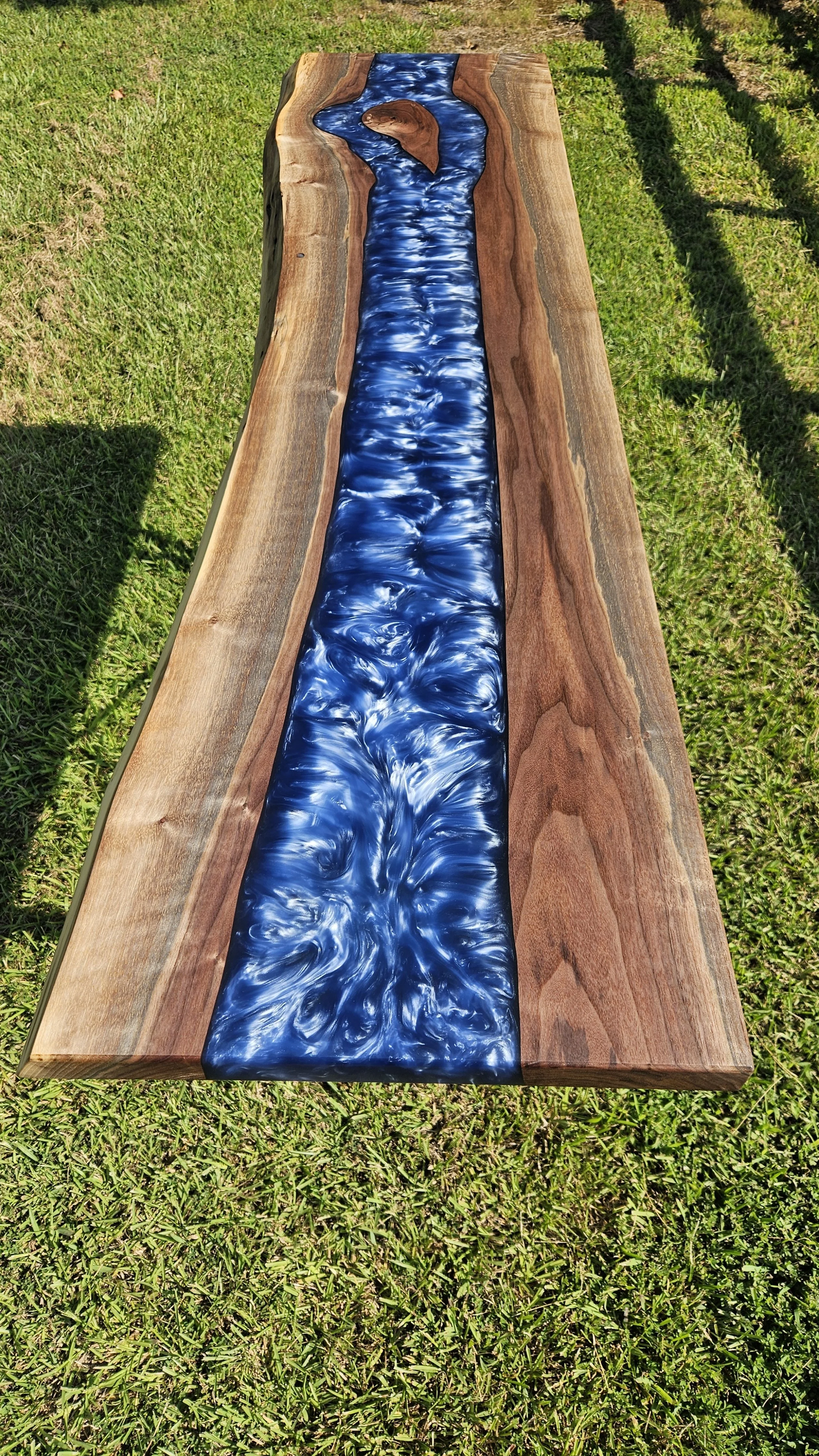 A wooden outdoor table with a blue and white resin river design in the center, placed on green grass.