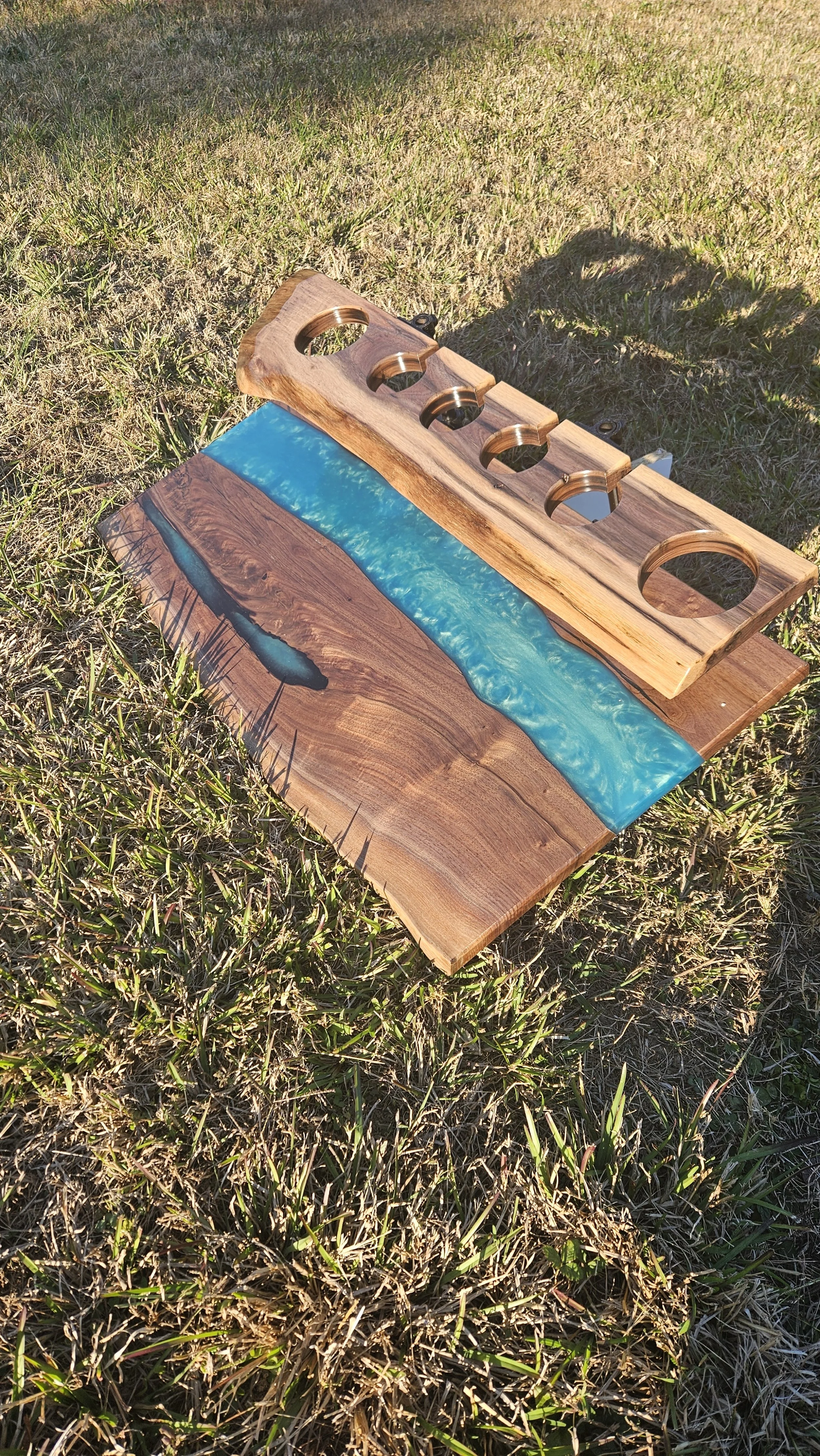 A wooden piece with circular cut-outs placed on a wooden board with a blue resin river, set on grass outdoors.